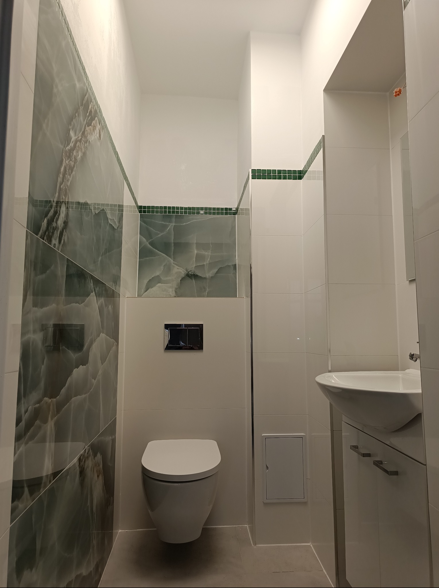 Nowoczesna łazienka z płytkami imitującymi marmur, wiszącą toaletą i umywalką z szafką. Ciemnozielony dekoracyjny pas płytek pod sufitem. Minimalistyczny styl.