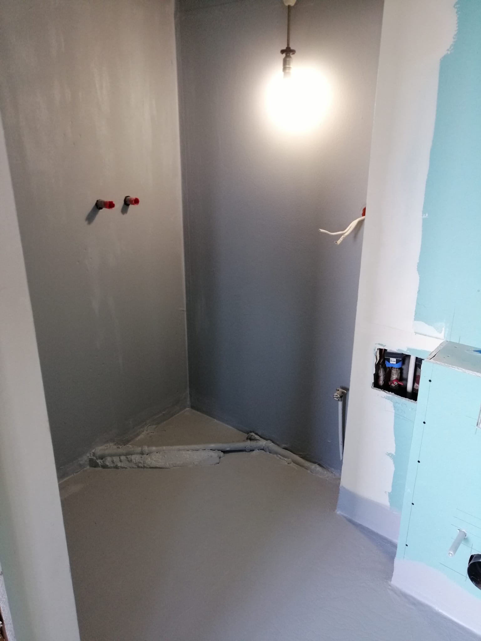 Łazienka w trakcie remontu: wylewka, ściany przygotowane do malowania, instalacja wodna. Widoczne rury i płyta karton-gips. Oświetlenie sufitowe.