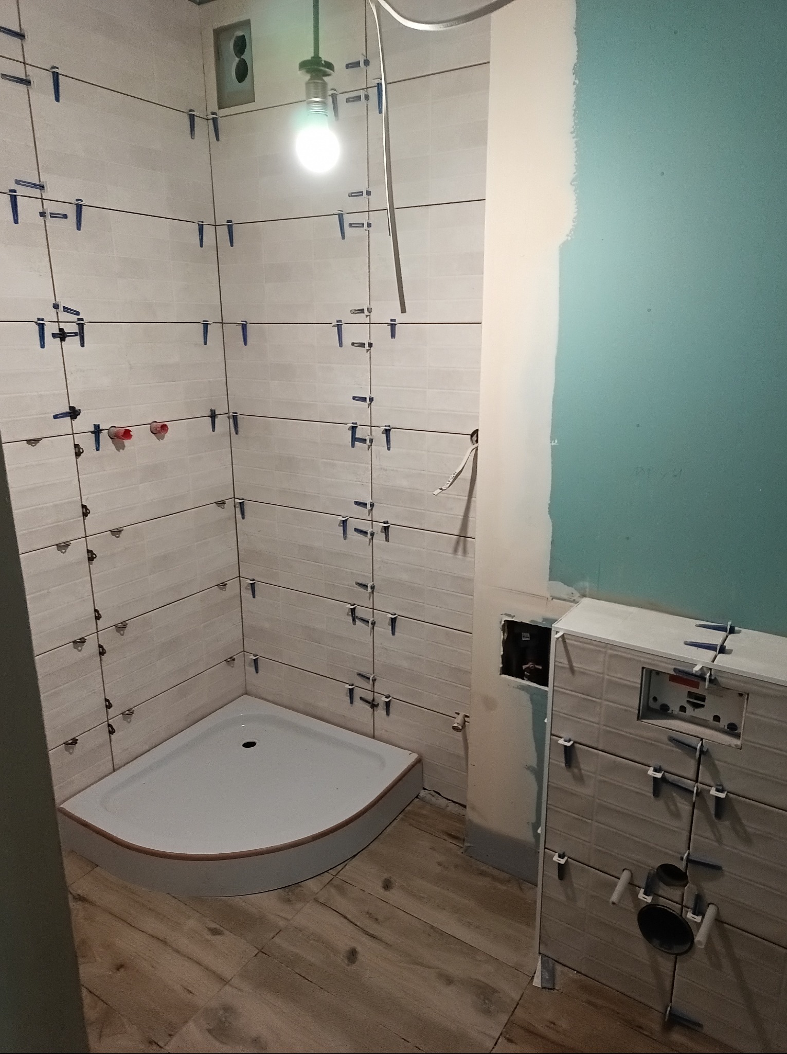 Białe płytki imitujące cegłę w trakcie montażu w łazience. Widoczny brodzik, stelaż podtynkowy i fragment ściany z płytą gipsową. Użyte niebieskie krzyżyki dystansowe.