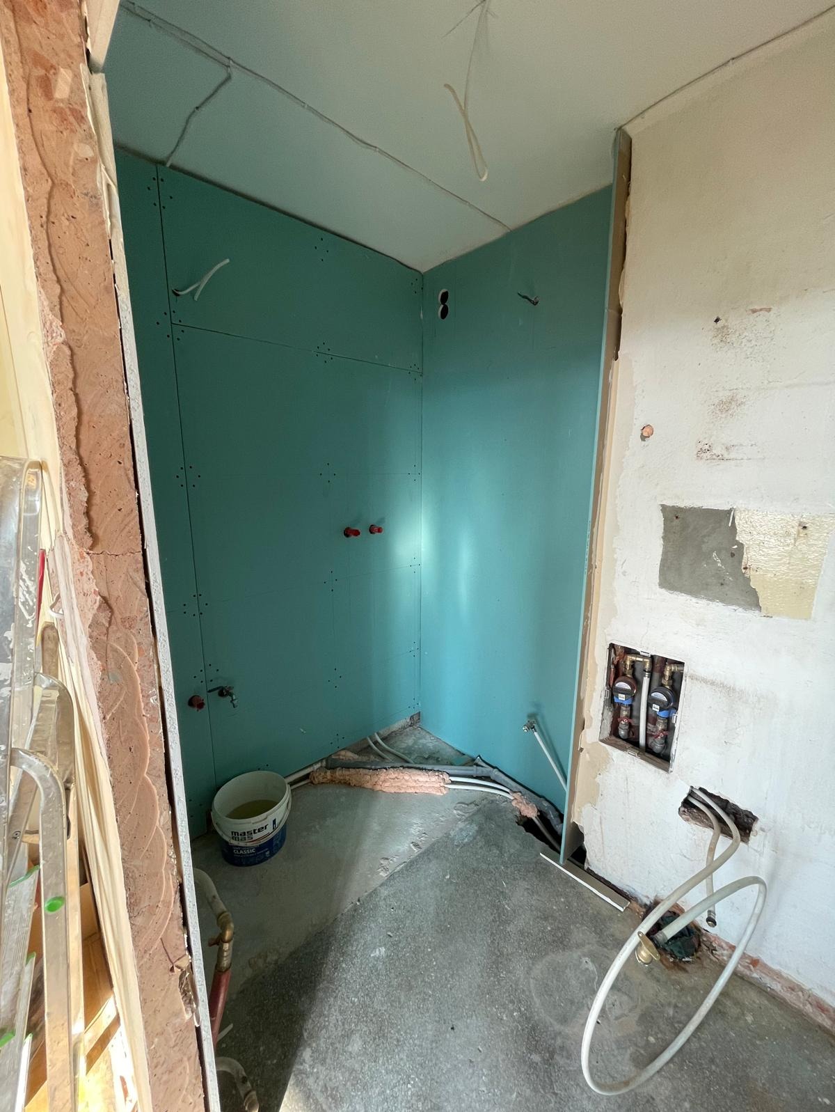 Narożnik łazienki w trakcie prac: zielone płyty gipsowe na ścianach, widoczne rury instalacji wodnej, fragment drabiny malarskiej. Etap przygotowania do dalszych prac.