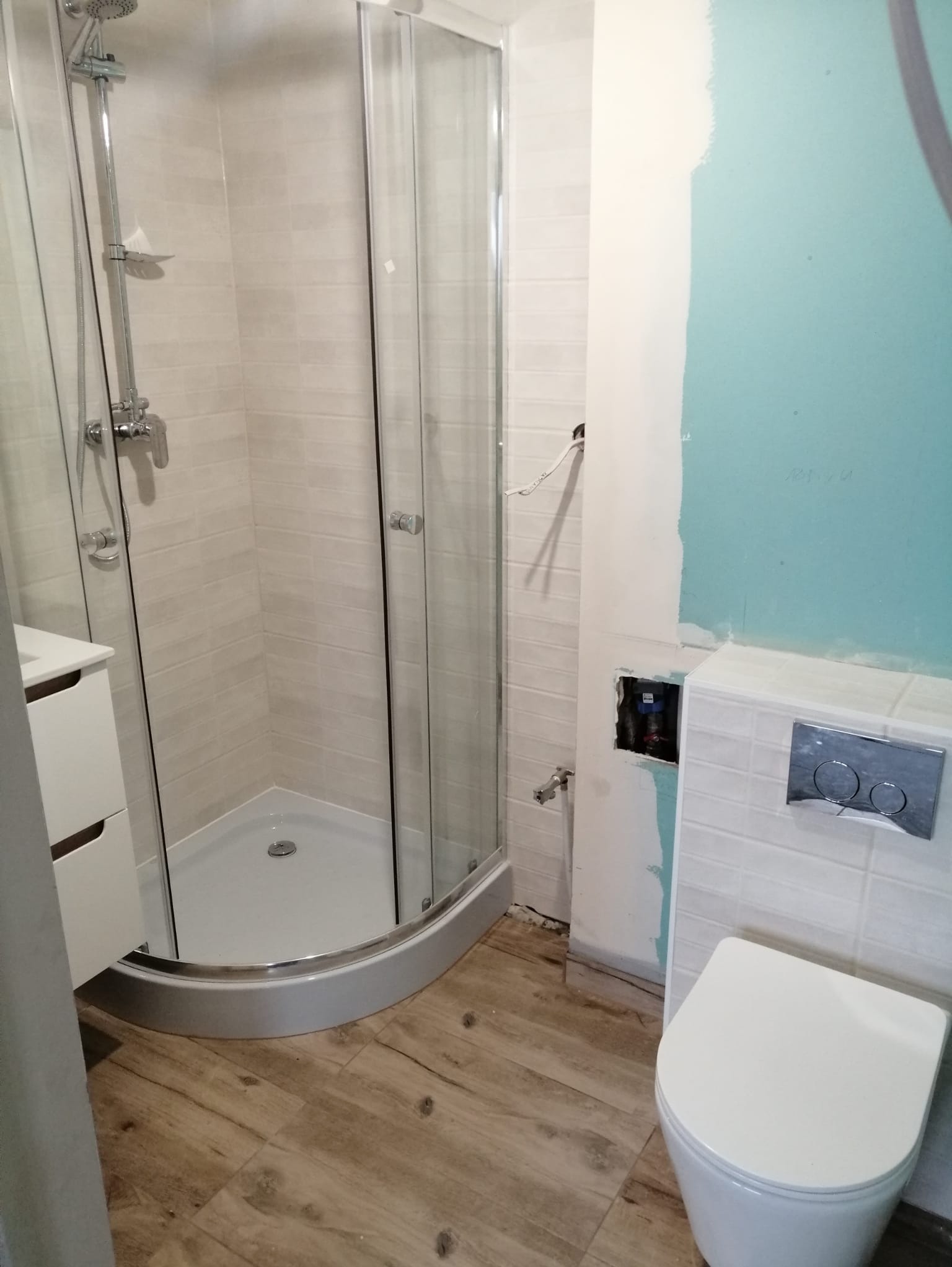 Narożna kabina prysznicowa z chromowaną armaturą, wisząca toaleta i drewnopodobna podłoga w trakcie prac wykończeniowych w łazience. Widoczne niedokończone ściany.