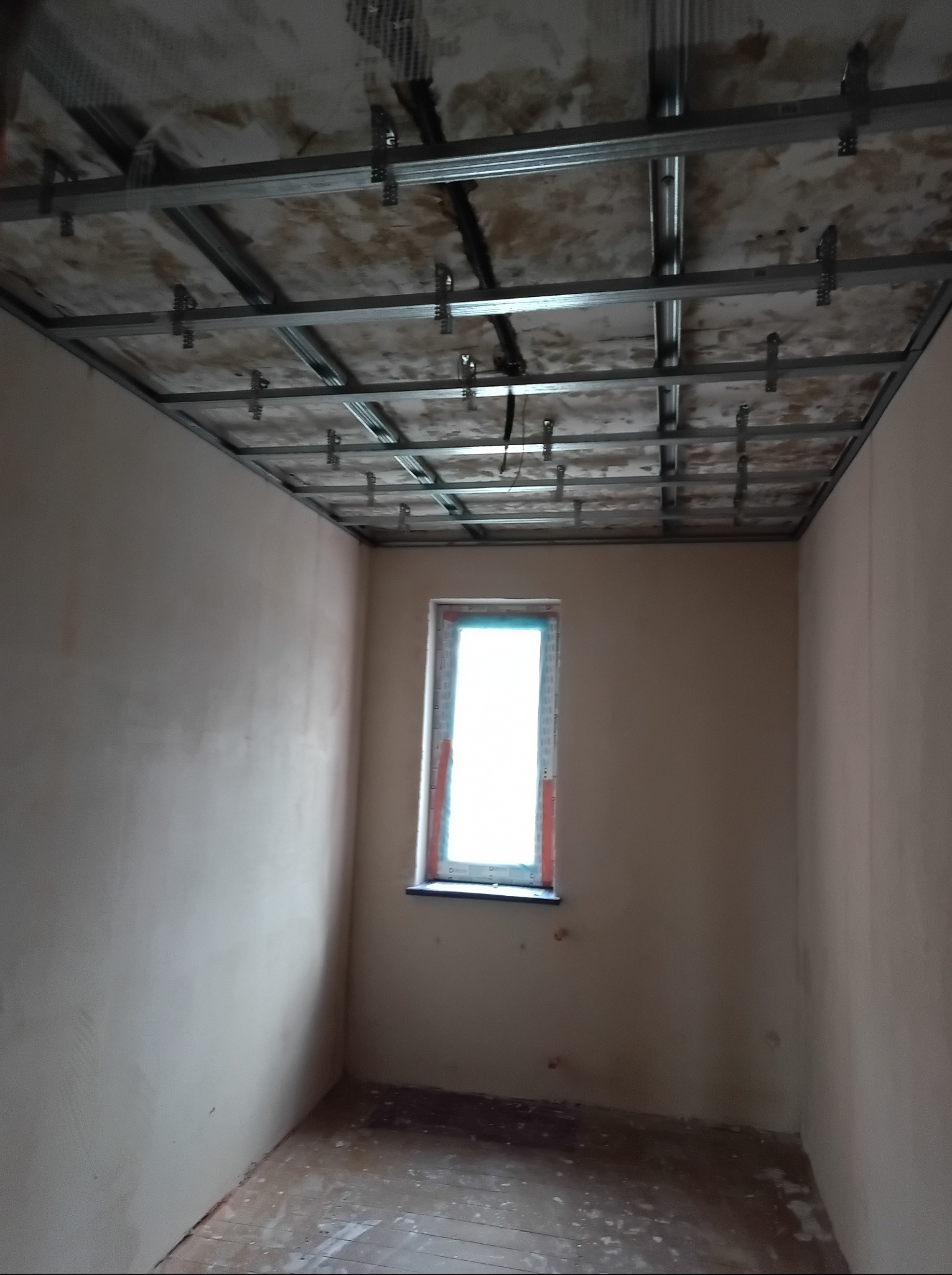 Pokój w trakcie montażu sufitu podwieszanego z metalowej konstrukcji. Widoczne profile i elementy mocujące. Ściany otynkowane, okno z widokiem na zewnątrz.