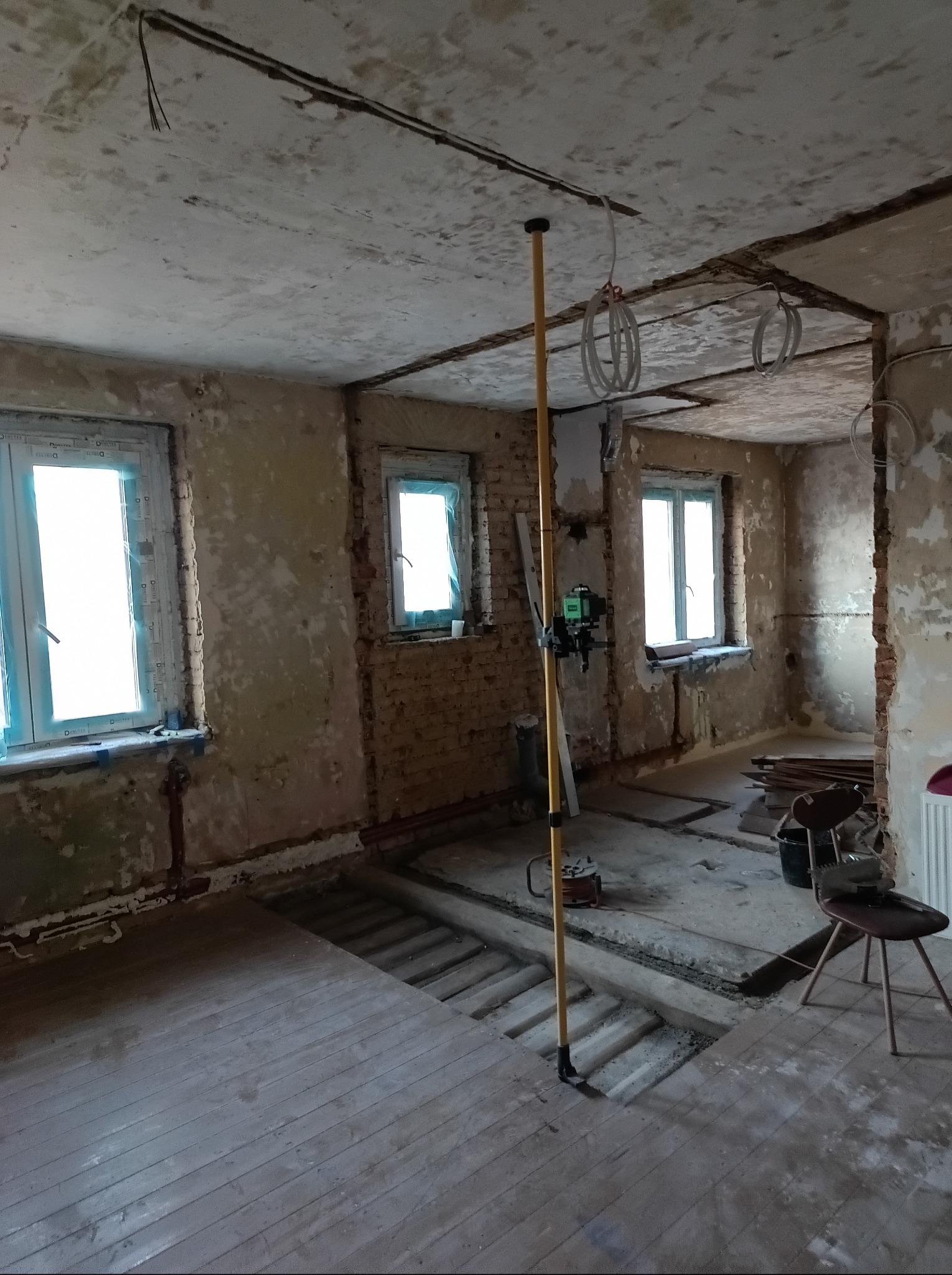Wnętrze w trakcie demontażu: odsłonięte cegły, resztki tynku, drewniana podłoga, widoczne instalacje elektryczne i poziomica laserowa na statywie.