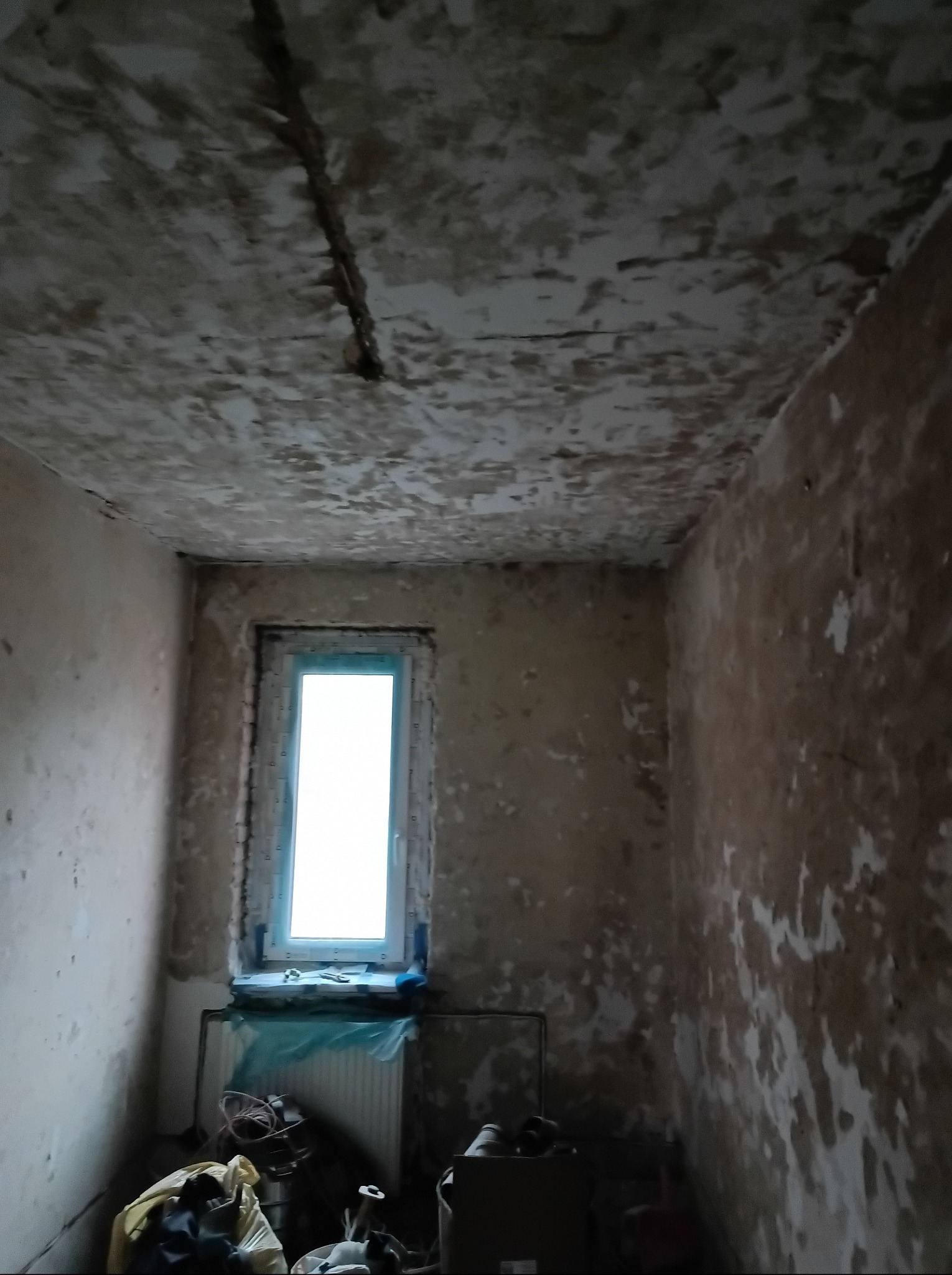 Pokój w trakcie przygotowań do remontu. Widoczne ściany ze śladami starej farby i gładzi, okno z grzejnikiem oraz narzędzia i materiały budowlane.