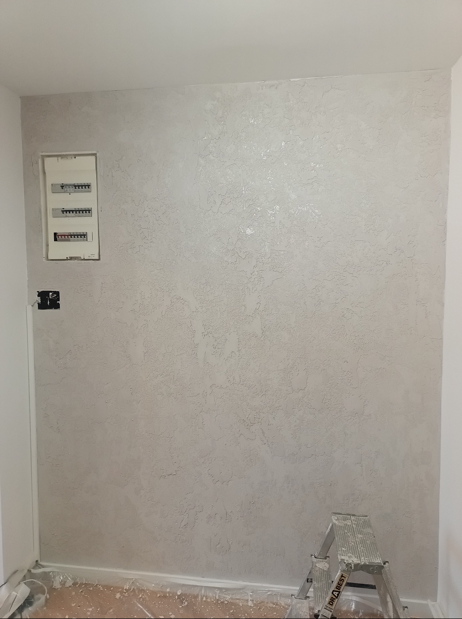 Dekoracyjna ściana po szpachlowaniu w Kłodzku z widoczną fakturą, zabezpieczona podłoga i drabina. Wykończenie wnętrza z detalami elektrycznymi.