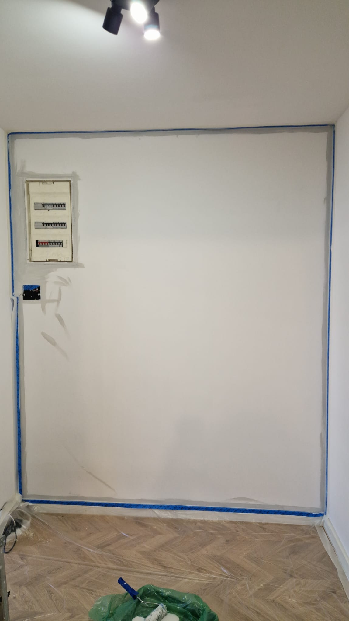 Biała ściana zabezpieczona niebieską taśmą malarską, przygotowana do malowania. Widoczna skrzynka elektryczna i folia ochronna na podłodze.