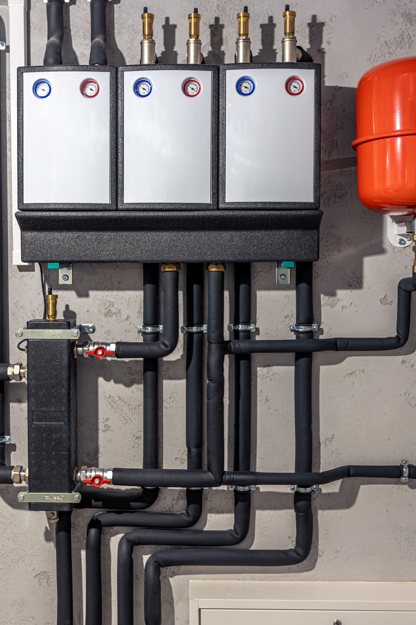 Złożona instalacja grzewcza z trzema panelami kontrolnymi, czarnymi rurami izolowanymi i czerwonym zbiornikiem wyrównawczym na tle ściany o szarej fakturze.