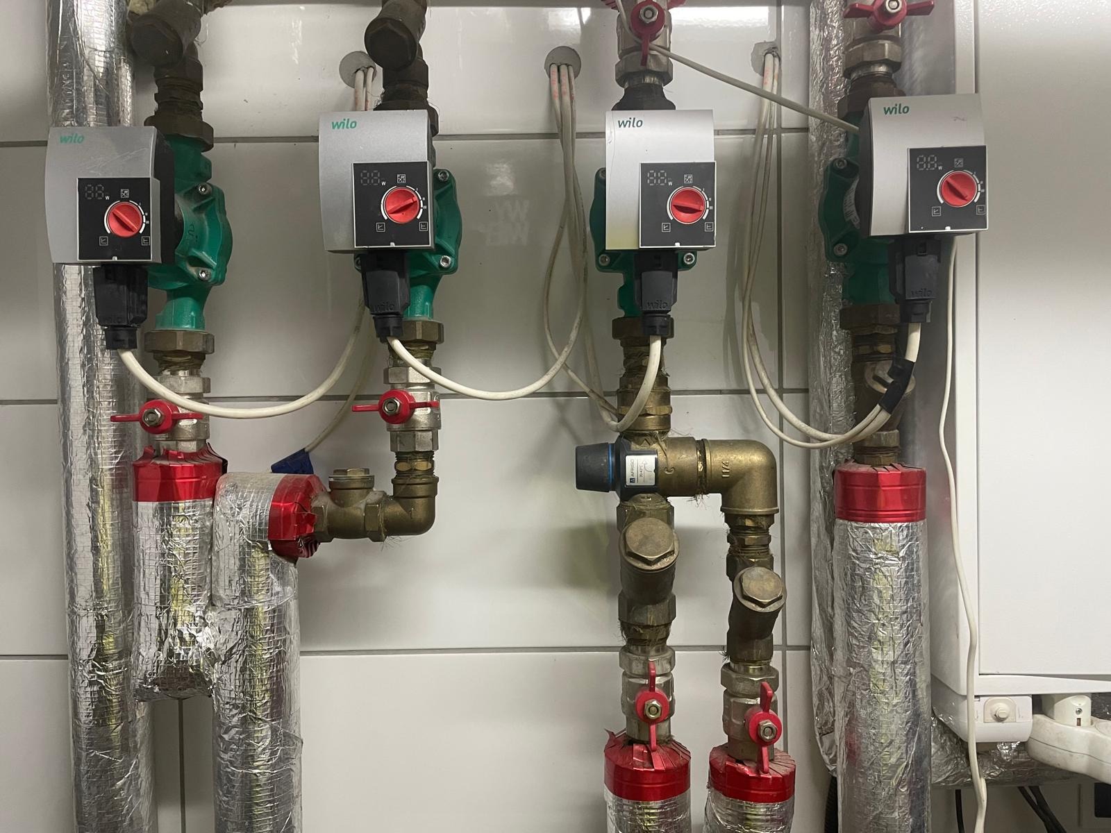 Trzy pompy obiegowe Wilo z zaworami i rurami, zamontowane na białej ścianie z płytek. Izolacja termiczna na rurach. Widoczne złącza i przewody.