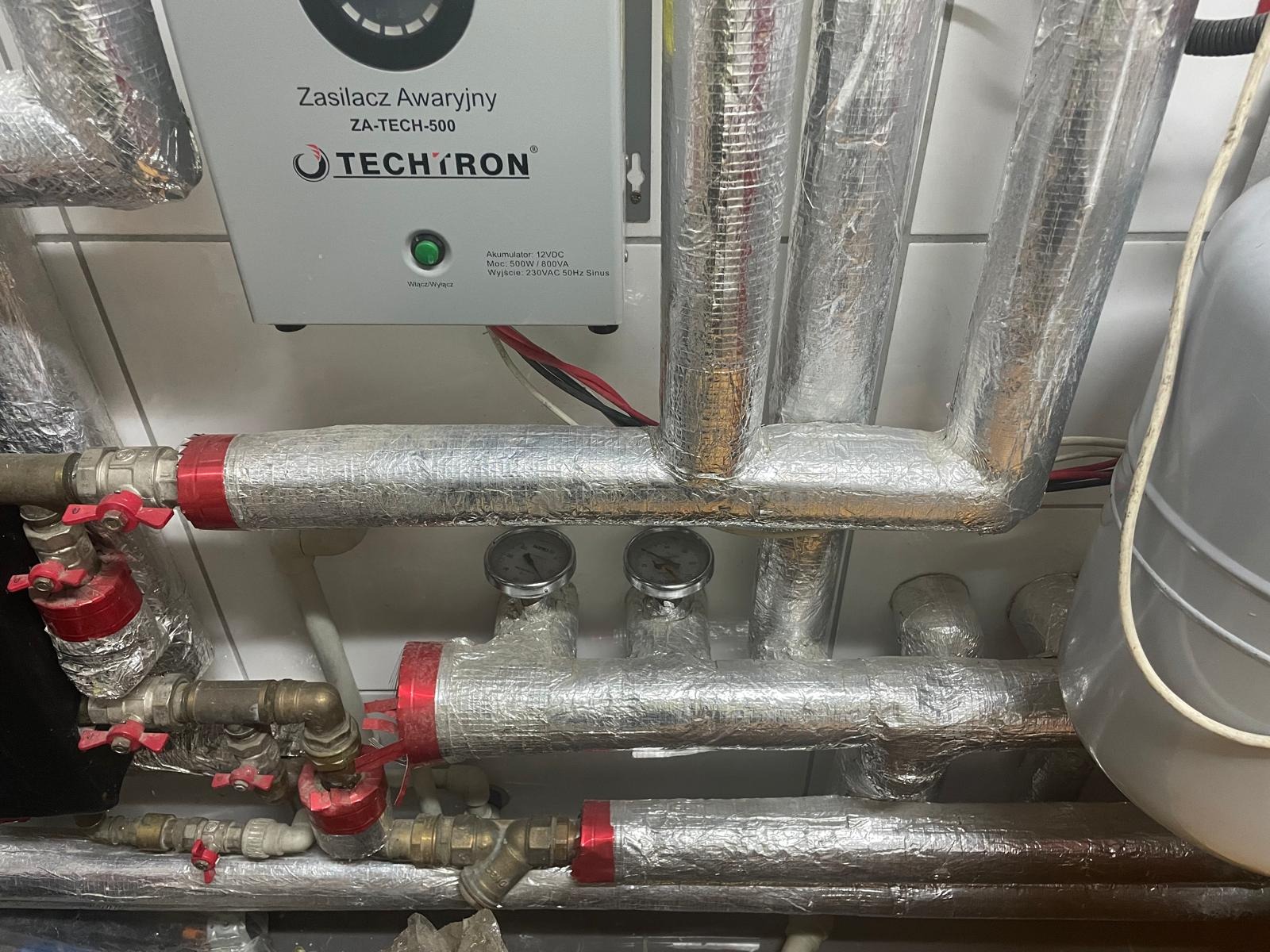 Izolowane rury instalacji grzewczej z zaworami i manometrami, zasilacz awaryjny Techtron ZA-TECH-500. Srebrna folia izolacyjna na rurach.
