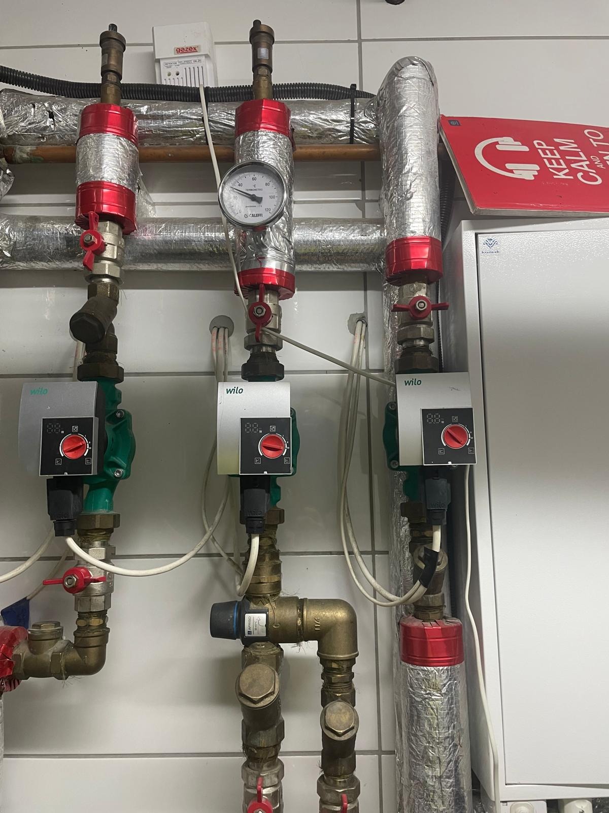 Instalacja grzewcza z pompami obiegowymi Wilo, rurami w izolacji i manometrem Caleffi. Widoczne zawory z czerwonymi pokrętłami i białe kable. Tło stanowią białe płytki.