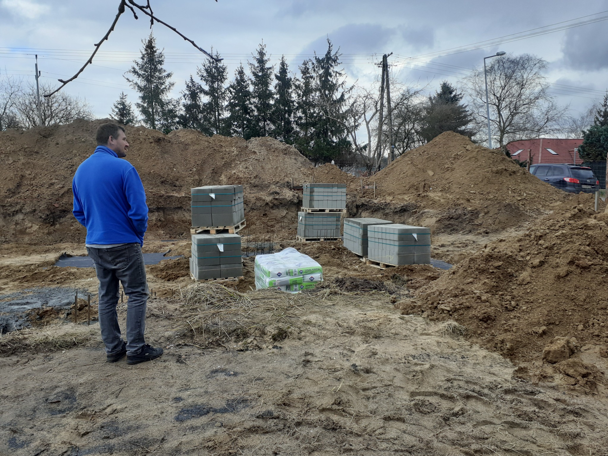 Układanie kostki brukowej: mężczyzna w niebieskiej bluzie obserwuje palety z kostką na placu budowy, w tle ziemia i drzewa. Widoczne przygotowania do prac brukarskich.