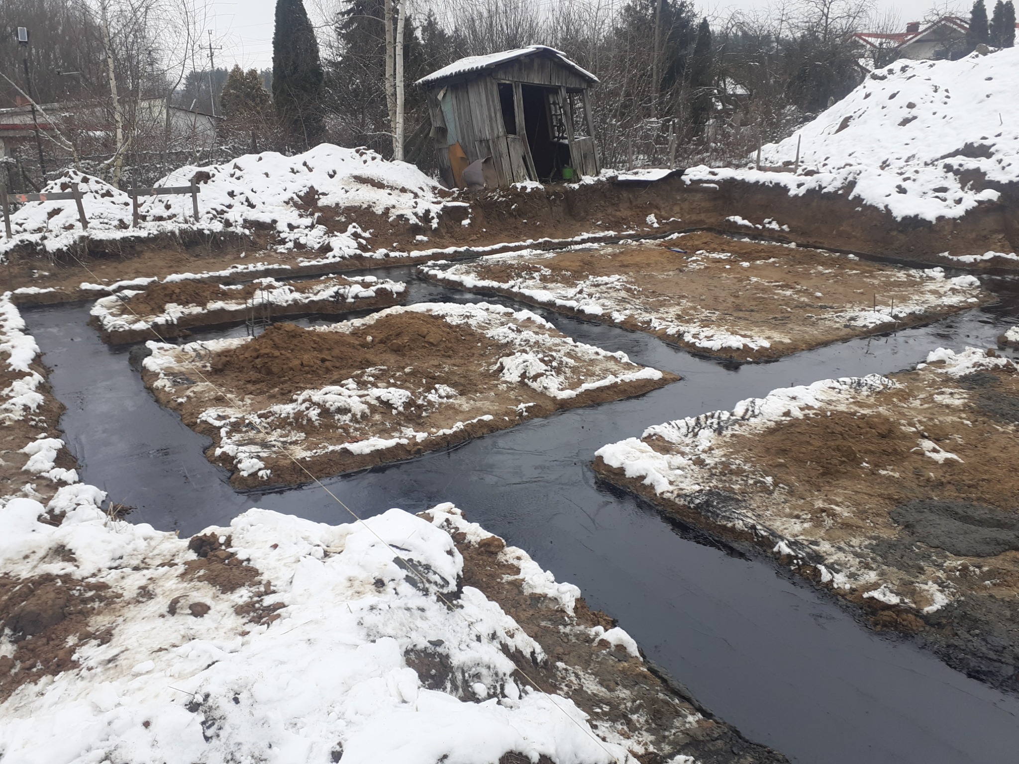 Przygotowanie fundamentów pod budynek w zimowej scenerii. Wykopane rowy zalane czarnym preparatem izolacyjnym, ziemia pokryta śniegiem. W tle drewniana szopa i drzewa.