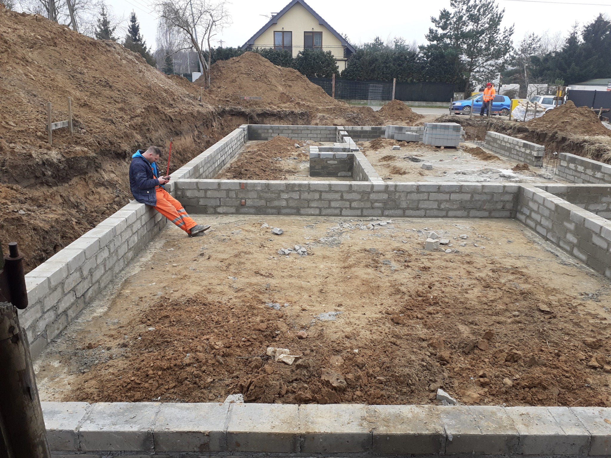 Fundamenty domu w trakcie budowy, z pracownikiem siedzącym na murze i korzystającym z telefonu. W tle widać wykopaną ziemię i częściowo ukończone ściany.