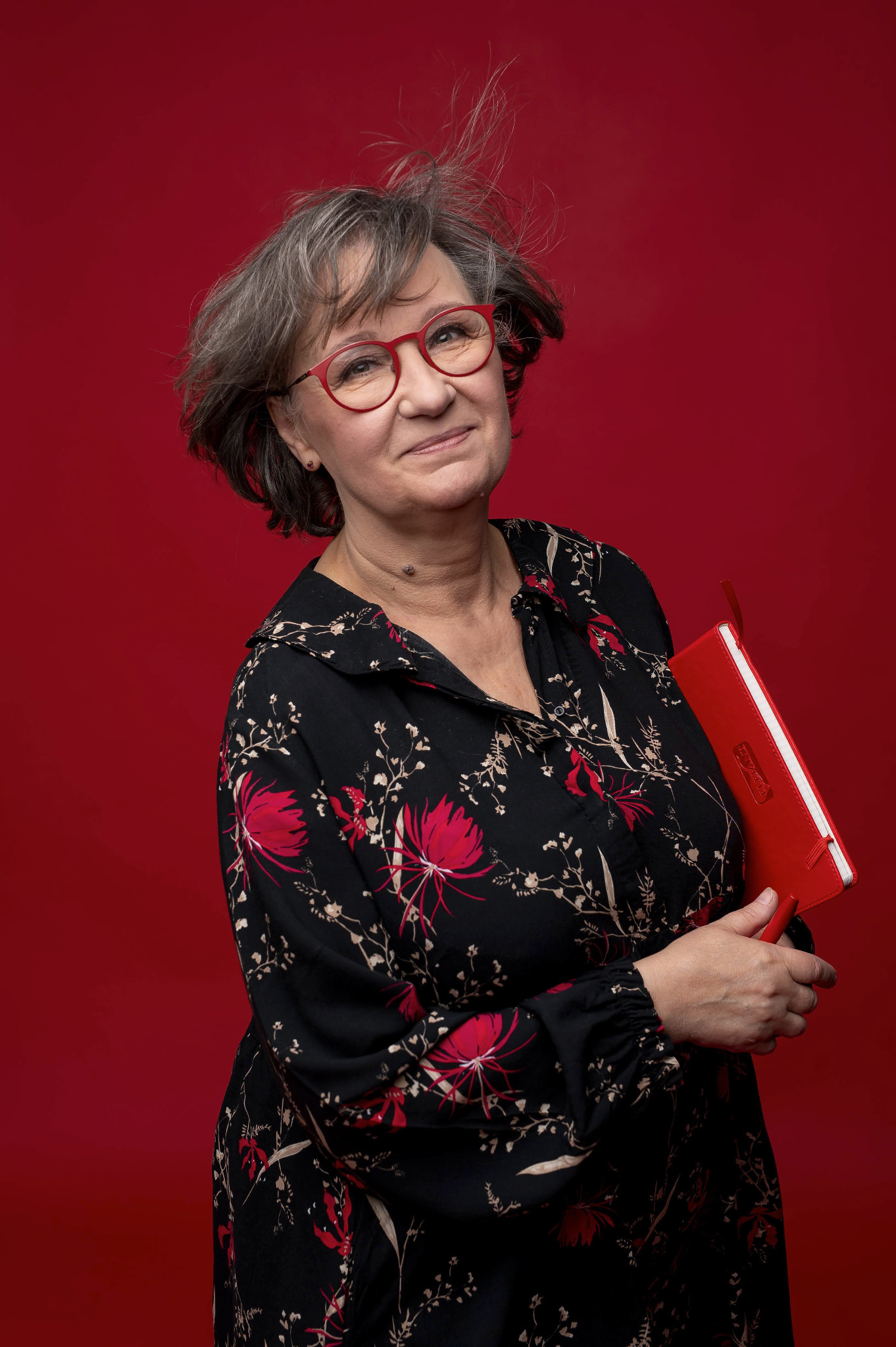 Uśmiechnięta kobieta w okularach z czerwonym notatnikiem na tle czerwonego studia. Włosy delikatnie rozwiane, wzrok skierowany w górę, sugerujący optymizm i świeże spojrzenie.