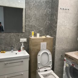 STAM - USŁUGI BUDOWŁANE - Wykończona łazienka z szarymi płytkami imitującymi marmur, białą szafką pod umywalką i toaletą myjącą. W tle widoczna pralka w opakowaniu.