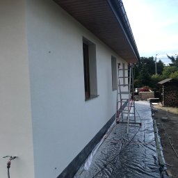 STAM - USŁUGI BUDOWŁANE - Ściana budynku po nałożeniu gładzi, widoczna drabina i zabezpieczona folią podstawa. Świeżo otynkowana elewacja w słoneczny dzień.