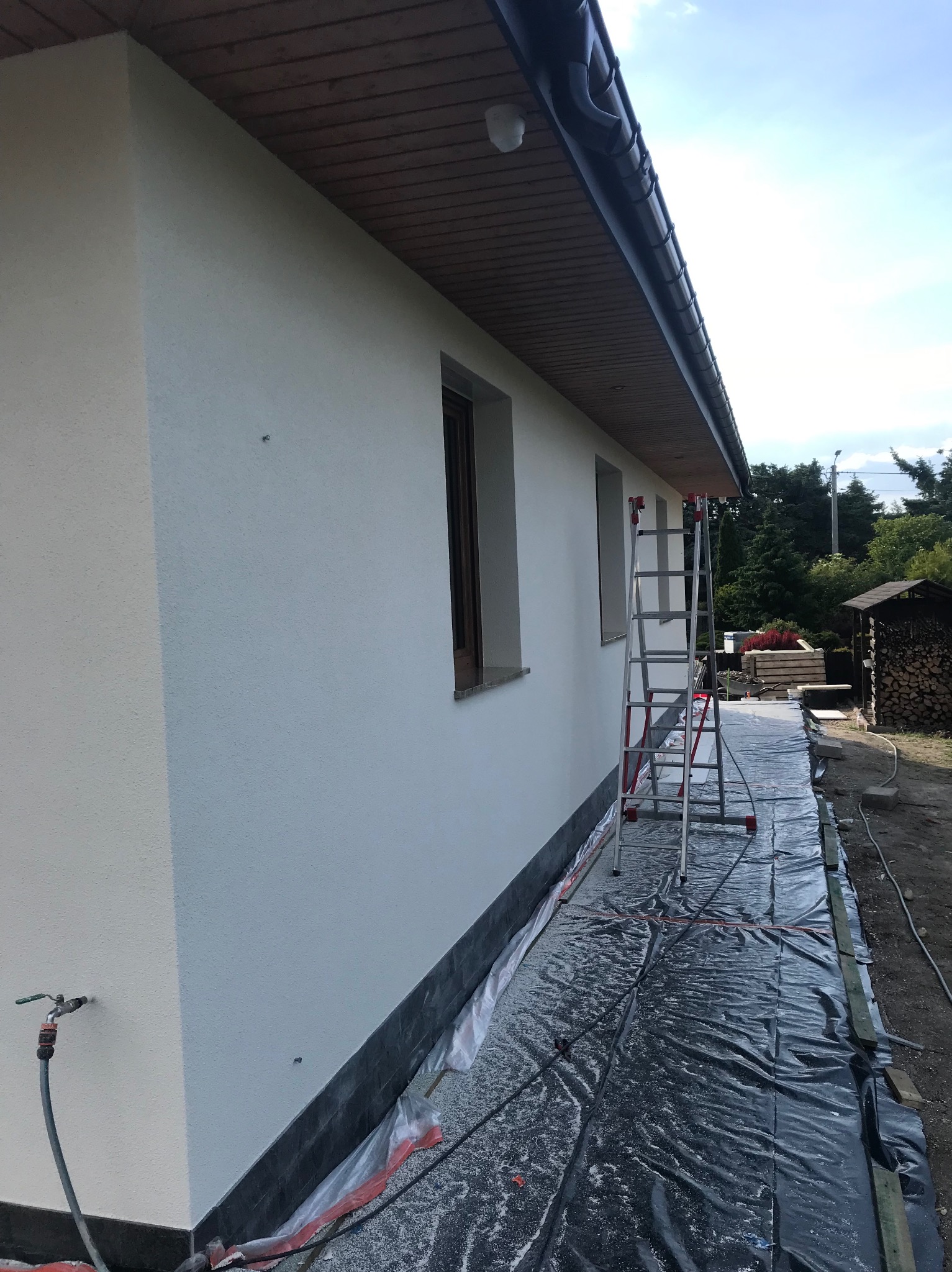 Ściana budynku po nałożeniu gładzi, widoczna drabina i zabezpieczona folią podstawa. Świeżo otynkowana elewacja w słoneczny dzień.