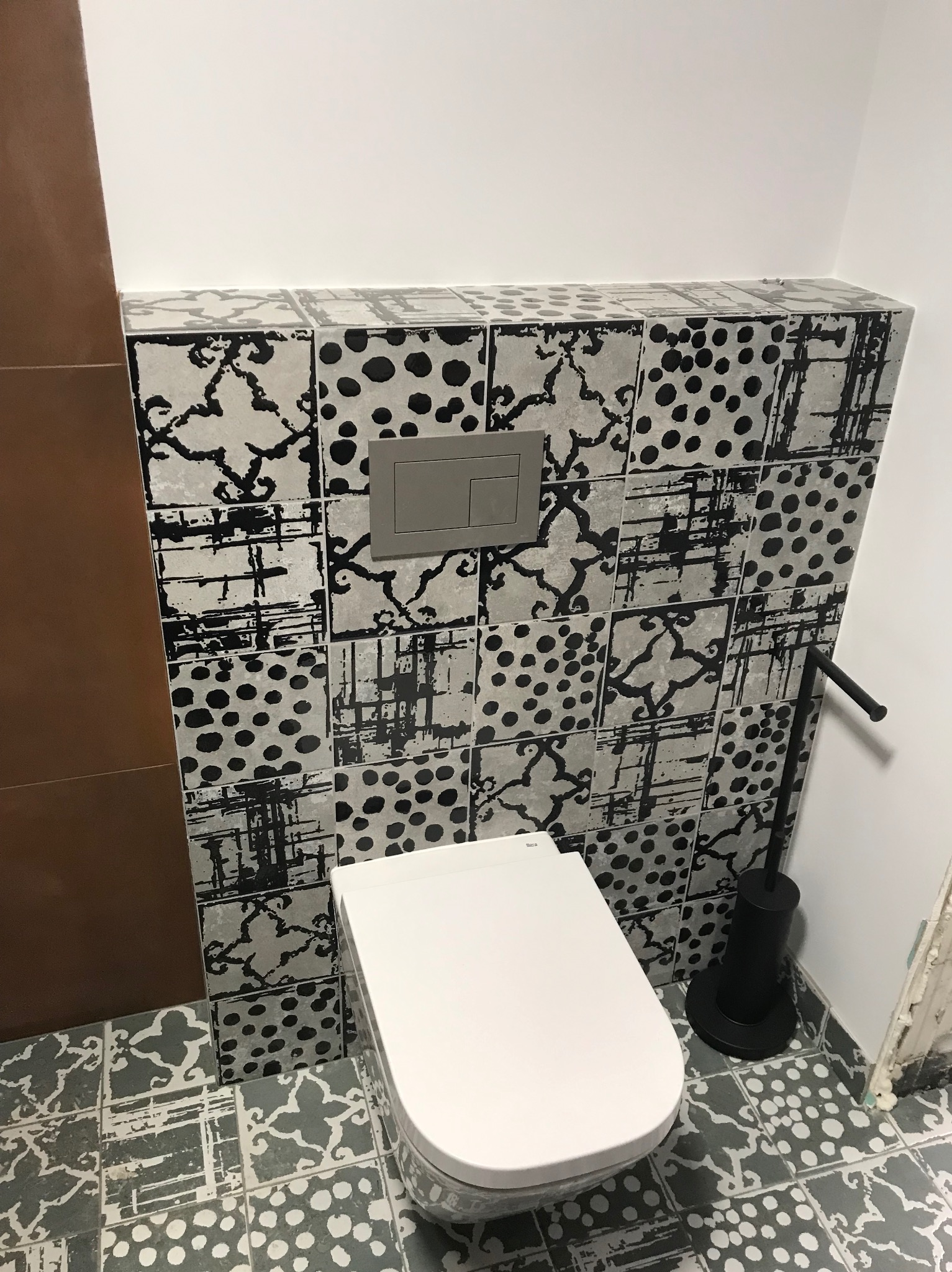 Nowoczesna toaleta podwieszana z szarą mozaiką w geometryczne wzory na ścianie i podłodze. Widoczny dozownik i szczotka do WC. Minimalistyczny design łazienki.