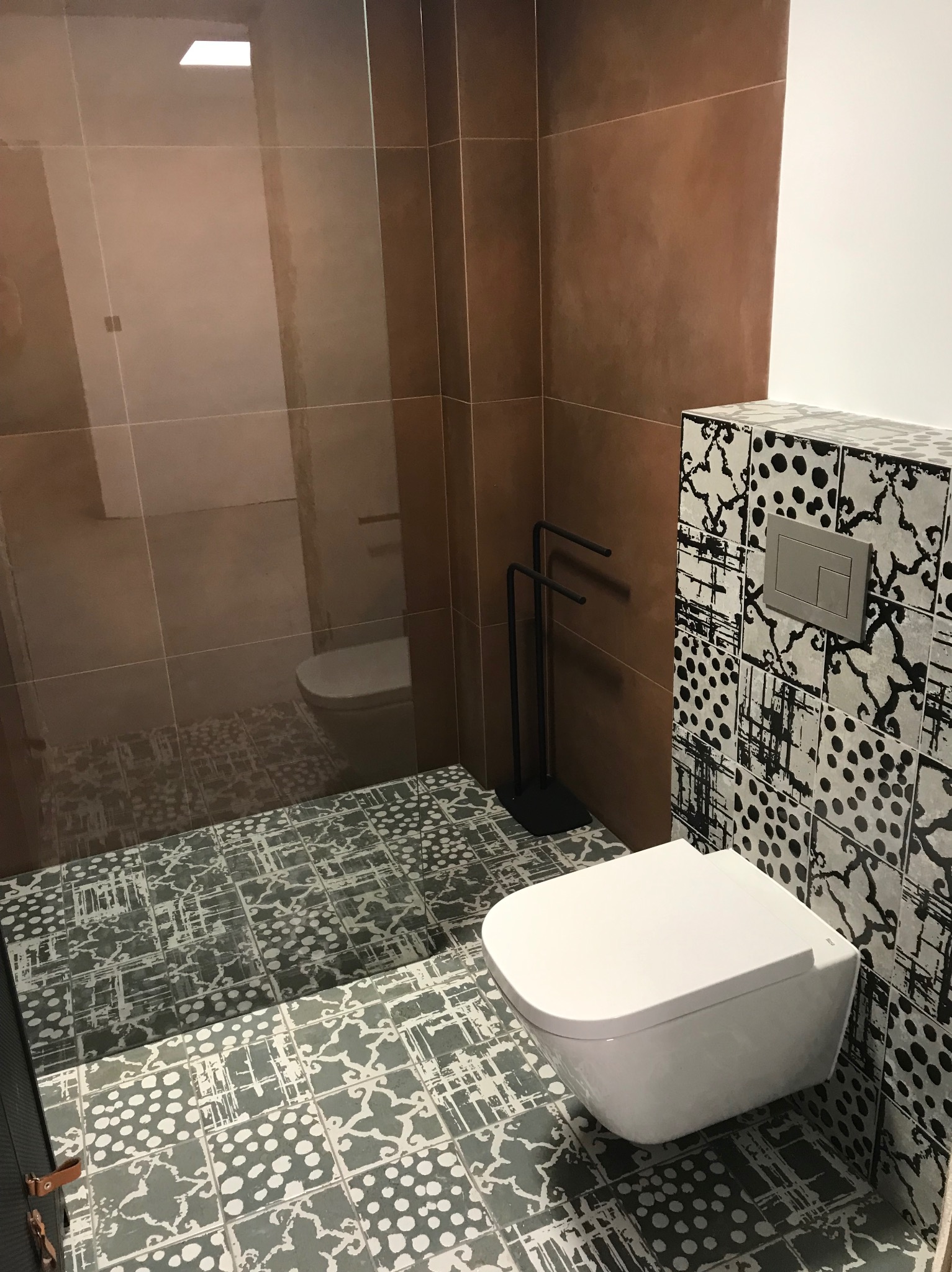 Nowoczesna łazienka z brązowymi płytkami, wzorzystą podłogą i wiszącą toaletą. Widoczne detale: czarny wieszak na ręczniki, geometryczny wzór na ścianie.