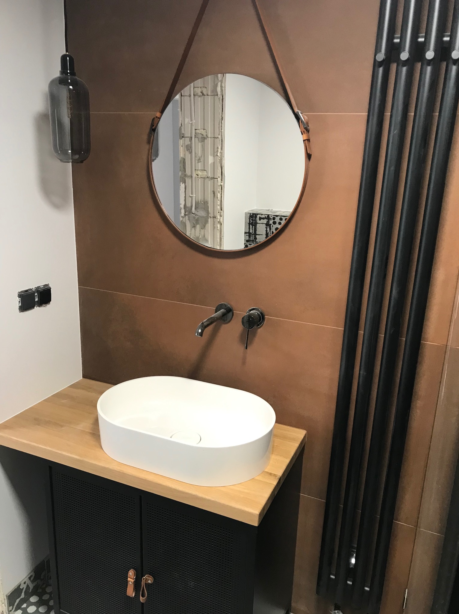 Nowoczesna łazienka z okrągłym lustrem na skórzanym pasku, białą umywalką nablatową i czarnym grzejnikiem drabinkowym na tle brązowych płytek.