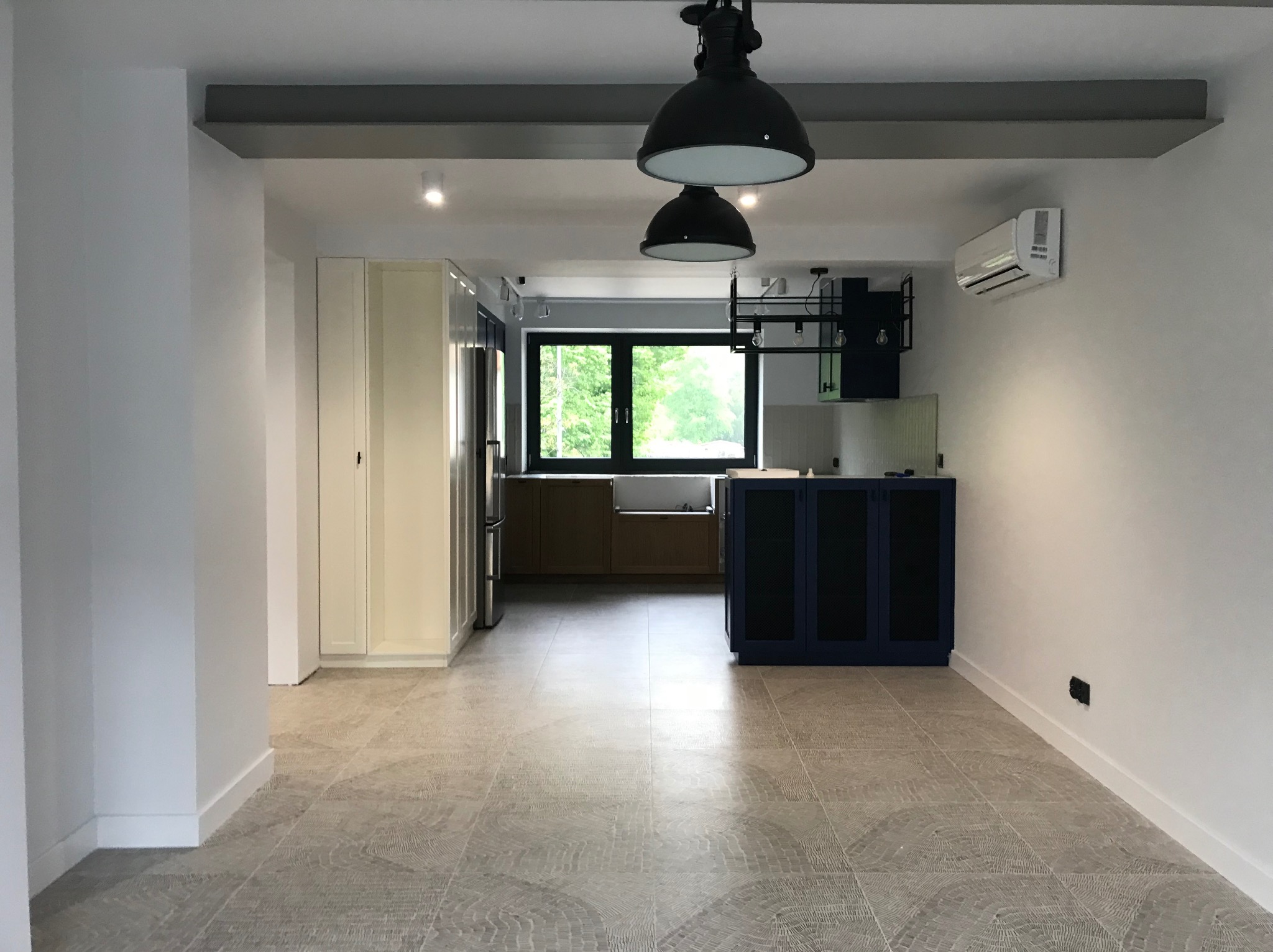 Przestronne wnętrze po remoncie: jasne ściany, wzorzysta podłoga, nowoczesne oświetlenie i widok na kuchnię z oknem. Minimalistyczny design z akcentami kolorystycznymi.