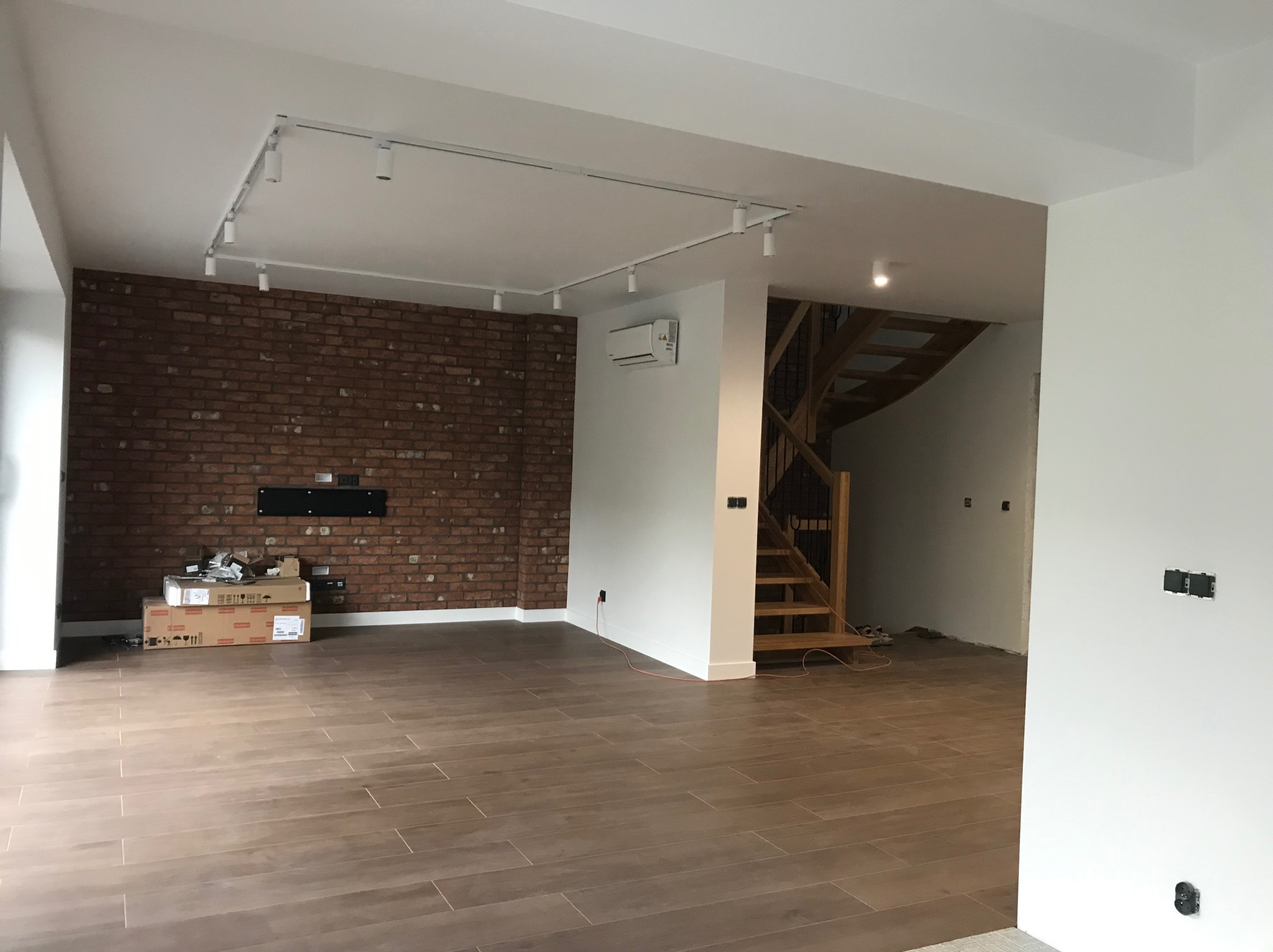 Przestronne wnętrze z brązową ceglaną ścianą, drewnianymi schodami i podłogą z płytek imitujących drewno. Białe ściany, sufit z oświetleniem szynowym i klimatyzacja.