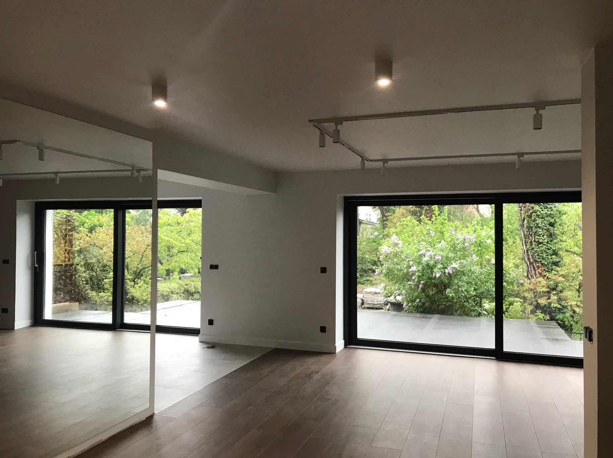 Jasne wnętrze z parkietem i dużymi oknami balkonowymi, widok na zieleń. Na suficie szyny z reflektorami. Część podłogi jeszcze nie wykończona.