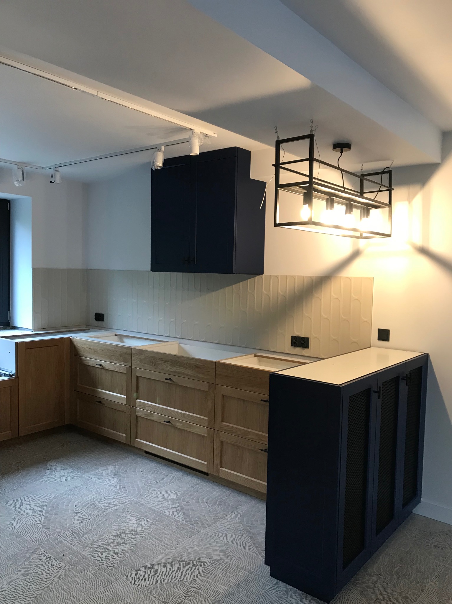 Montaż kuchni z drewnianymi szafkami i ciemnymi elementami, oświetleniem sufitowym i wzorzystą podłogą. Widoczne płytki na ścianie.