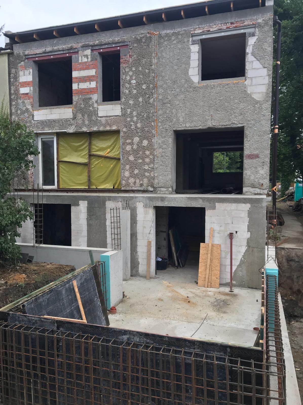 Surowy stan dwukondygnacyjnego budynku w trakcie remontu: odsłonięte cegły, okna zabite folią, widoczne zbrojenia i betonowe elementy konstrukcyjne.