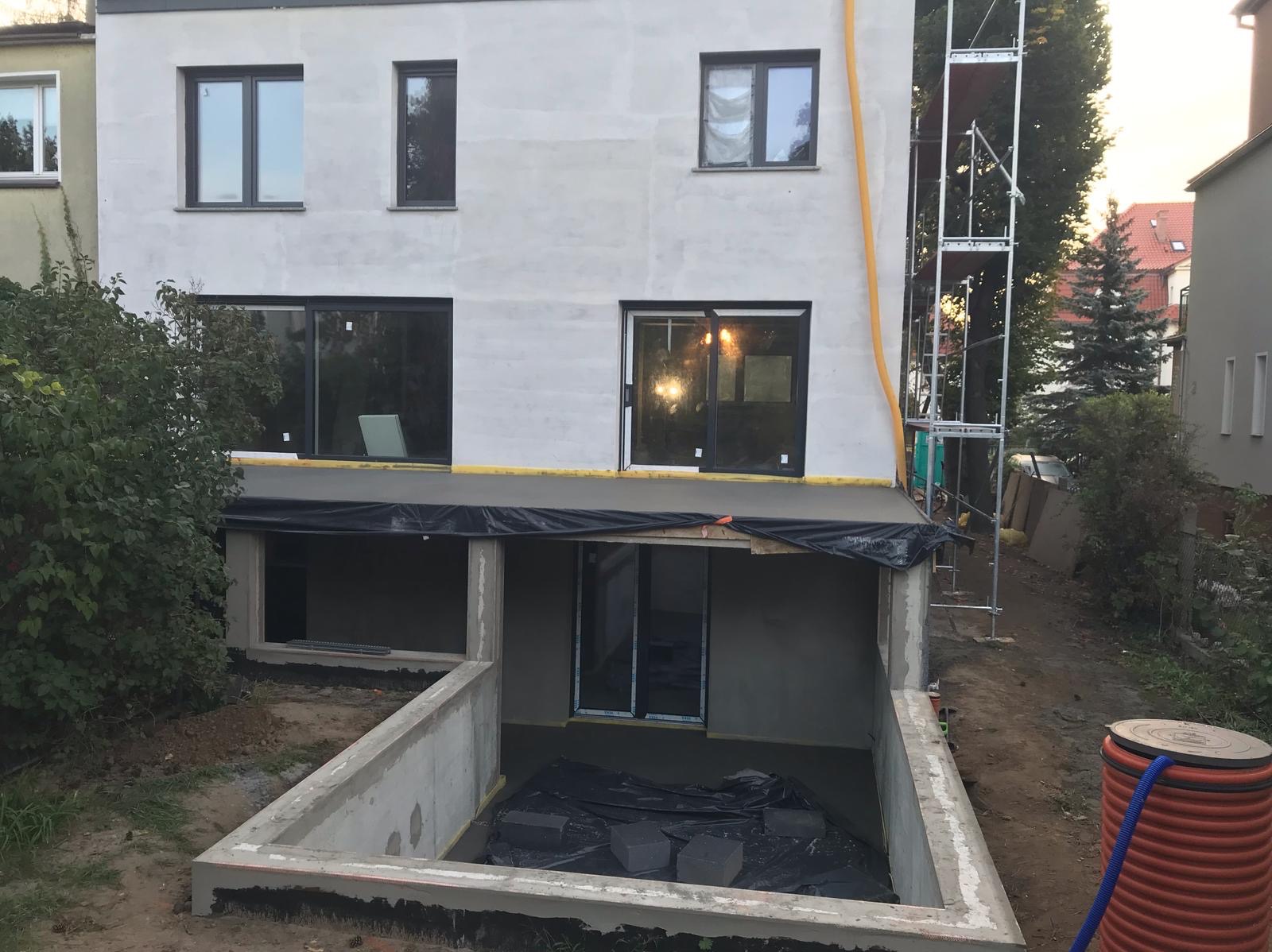 Budynek w trakcie prac wykończeniowych: ocieplenie fundamentów, widoczne rusztowania i przygotowane okna. Prace ziemne wokół budynku.