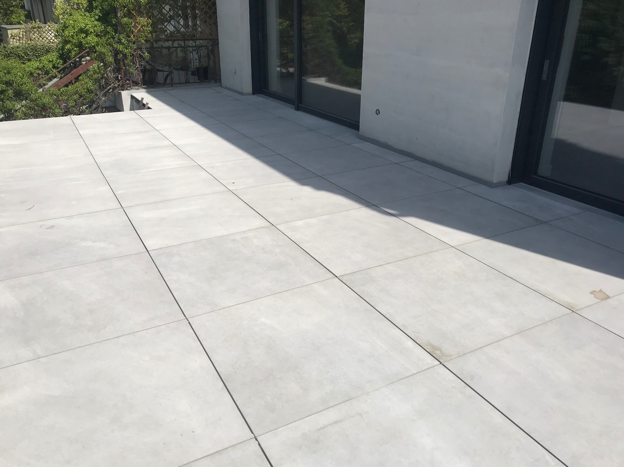 Jasnoszara, nowoczesna podłoga z płytek na balkonie, z widocznymi drzwiami balkonowymi i zielenią w tle. Minimalistyczny design, równomierne oświetlenie.