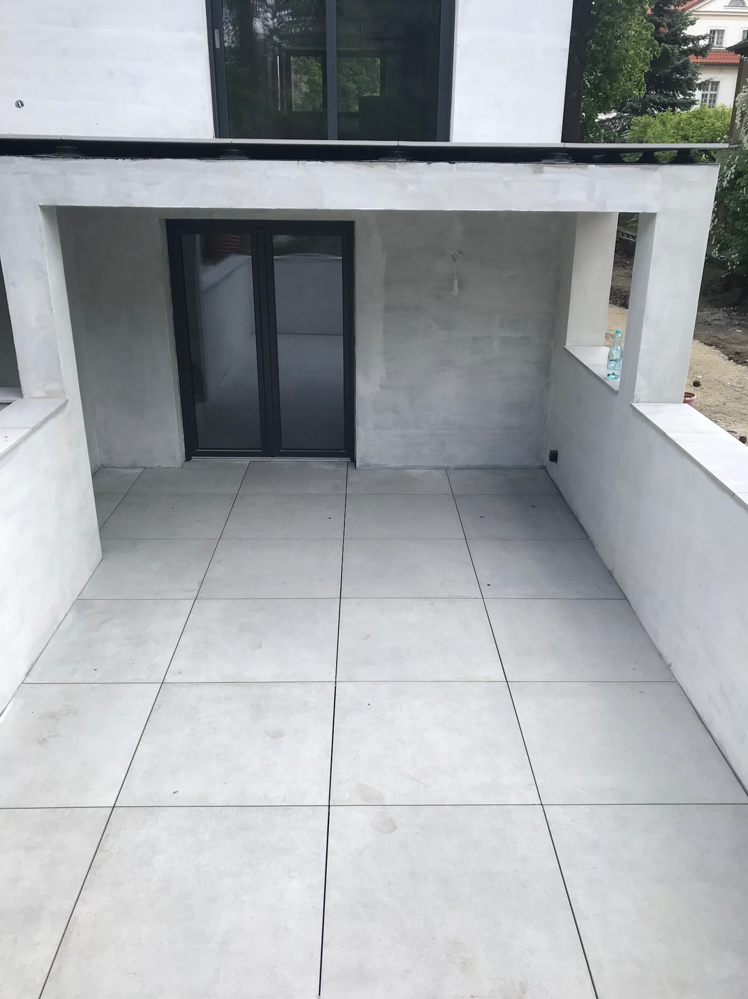 Wykończony balkon z szarymi płytkami i świeżo pomalowanymi ścianami. Widoczna butelka wody na parapecie. Minimalistyczny design.