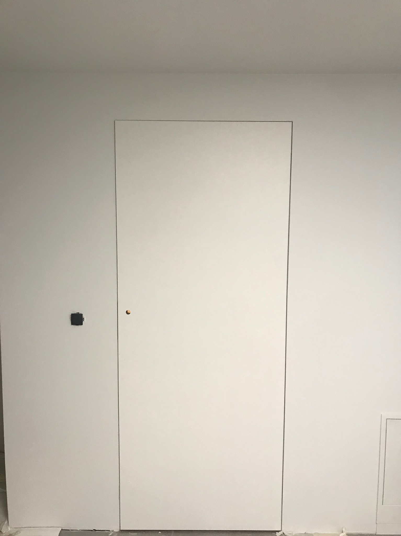 Minimalistyczne, białe drzwi ukryte w ścianie, z widocznym otworem na klamkę i niezamaskowanym gniazdkiem elektrycznym po lewej stronie.