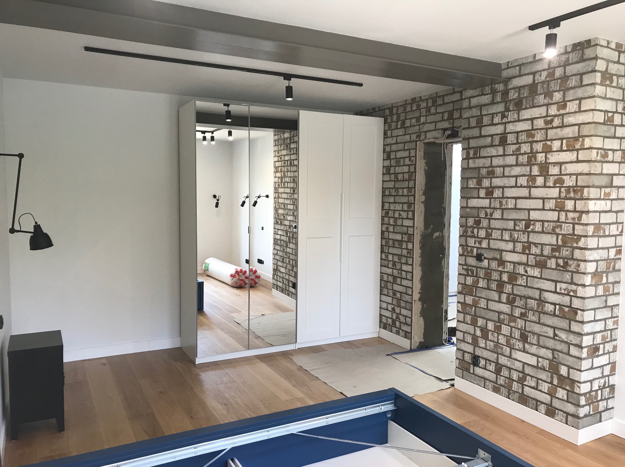 Pokój w trakcie wykańczania: drewniana podłoga, ceglana ściana z surowym otworem drzwiowym, biała szafa z lustrem i rolka materiału w odbiciu.