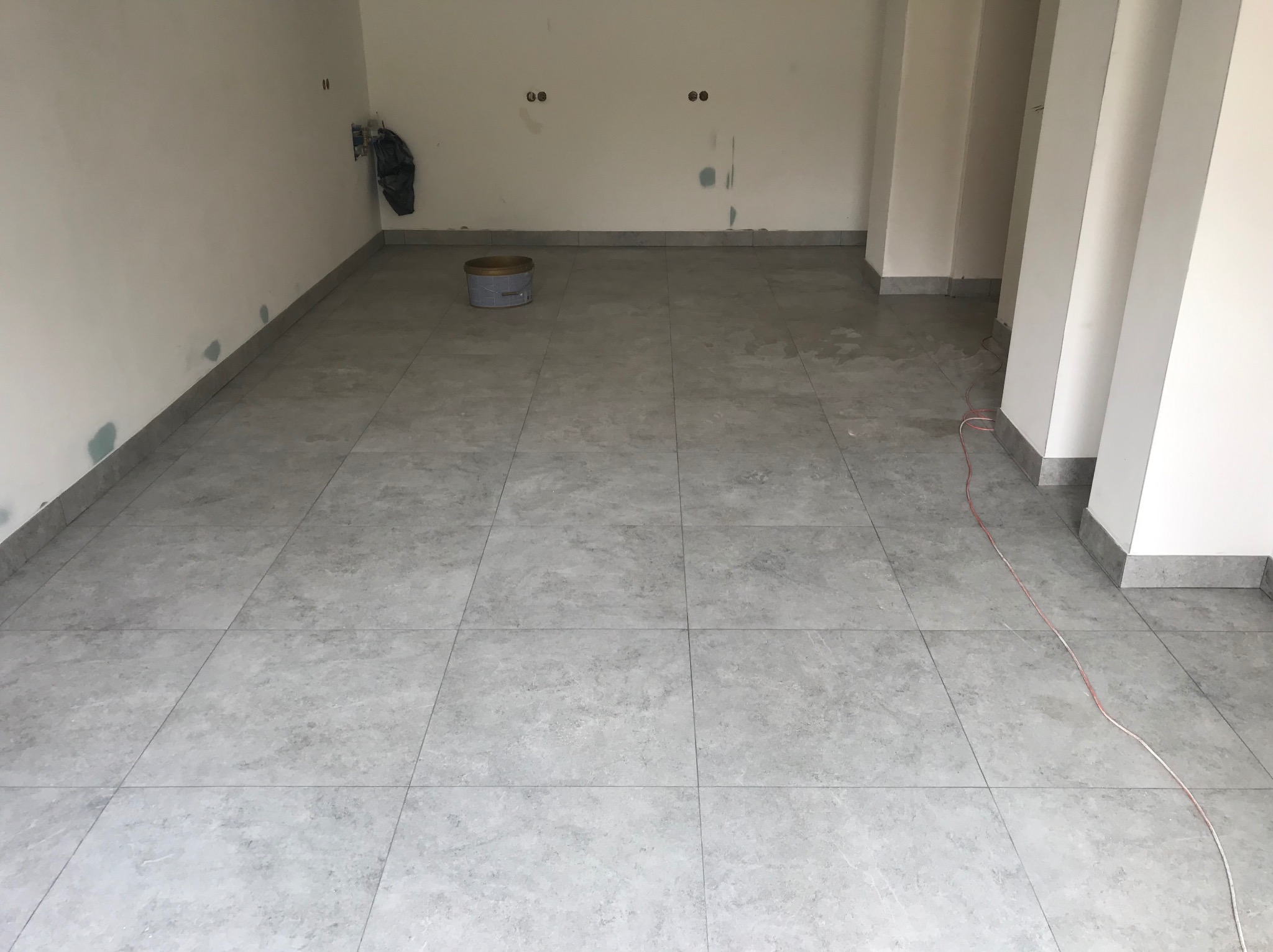 Wykończona podłoga z szarych płytek w pomieszczeniu w Świdnicy. Widoczny fragment ściany z puszkami elektrycznymi i wiadro z farbą. Minimalistyczny styl.