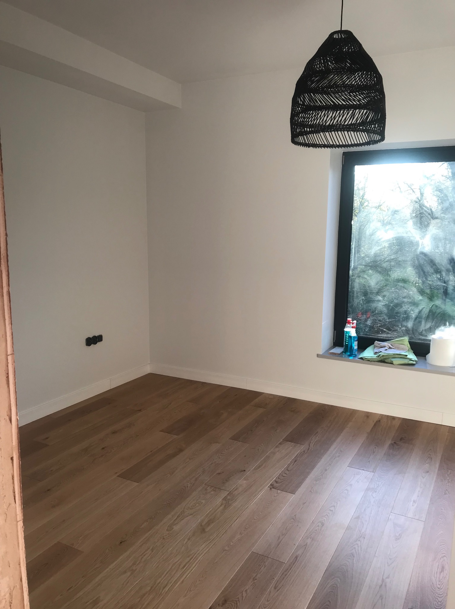 Jasne panele podłogowe w nowym pokoju z białymi ścianami, czarnym oknem i lampą. Widoczne gniazdka i środki czystości na parapecie. Wnętrze po remoncie.