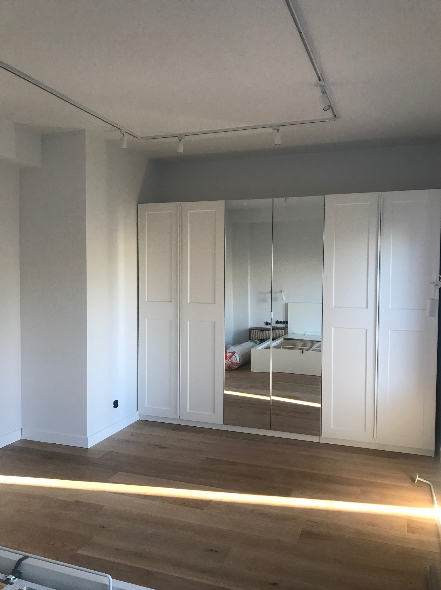 Jasne wnętrze z drewnianą podłogą i białym, czterodrzwiowym lustrzanym szafą wnękową pod sufitem z szynoprzewodem oświetleniowym.