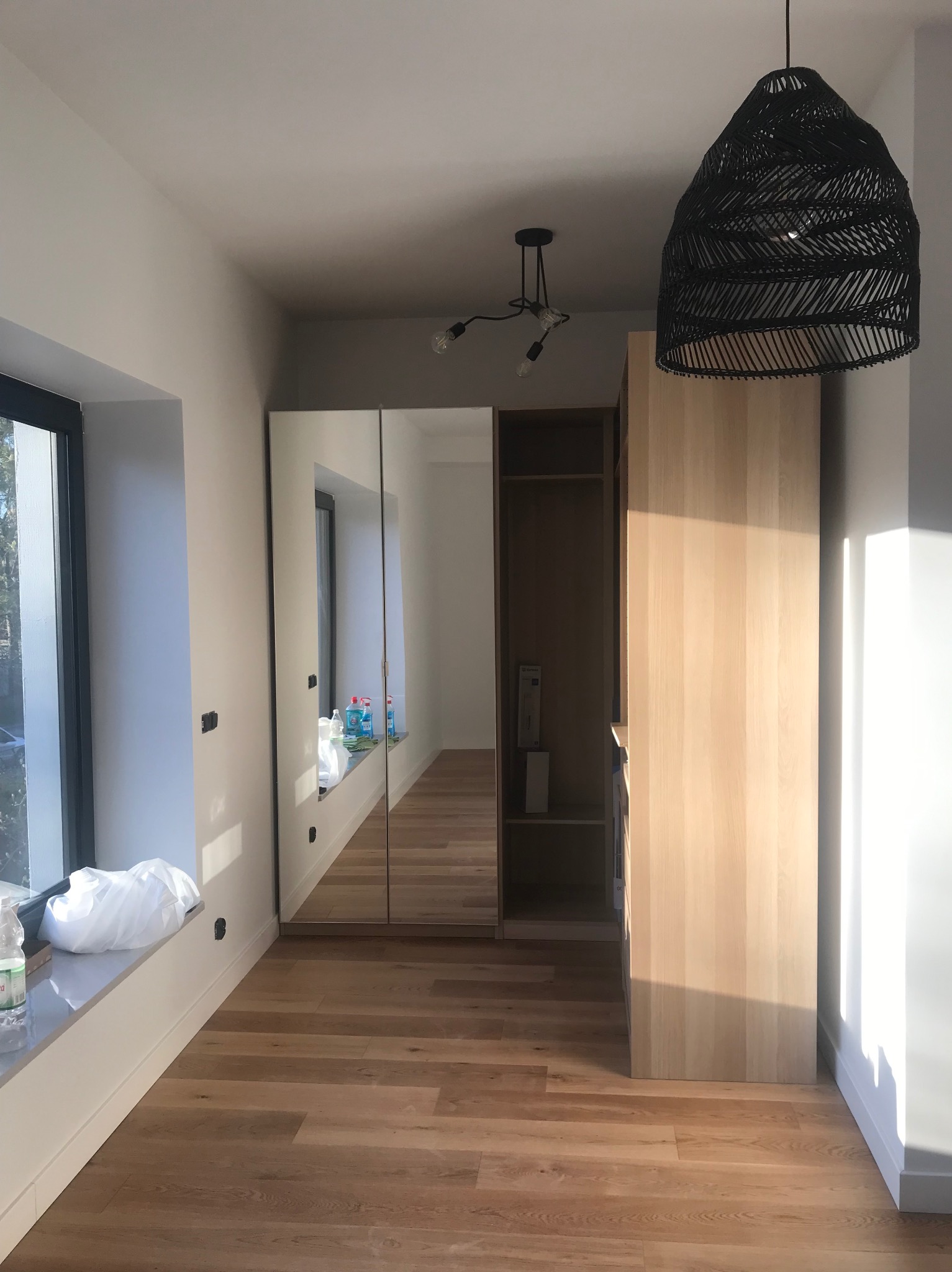 Jasne wnętrze z drewnianą podłogą i szafą wnękową z lustrzanymi drzwiami. Widoczne okno, lampa wisząca i akcesoria na parapecie. Minimalistyczny design.