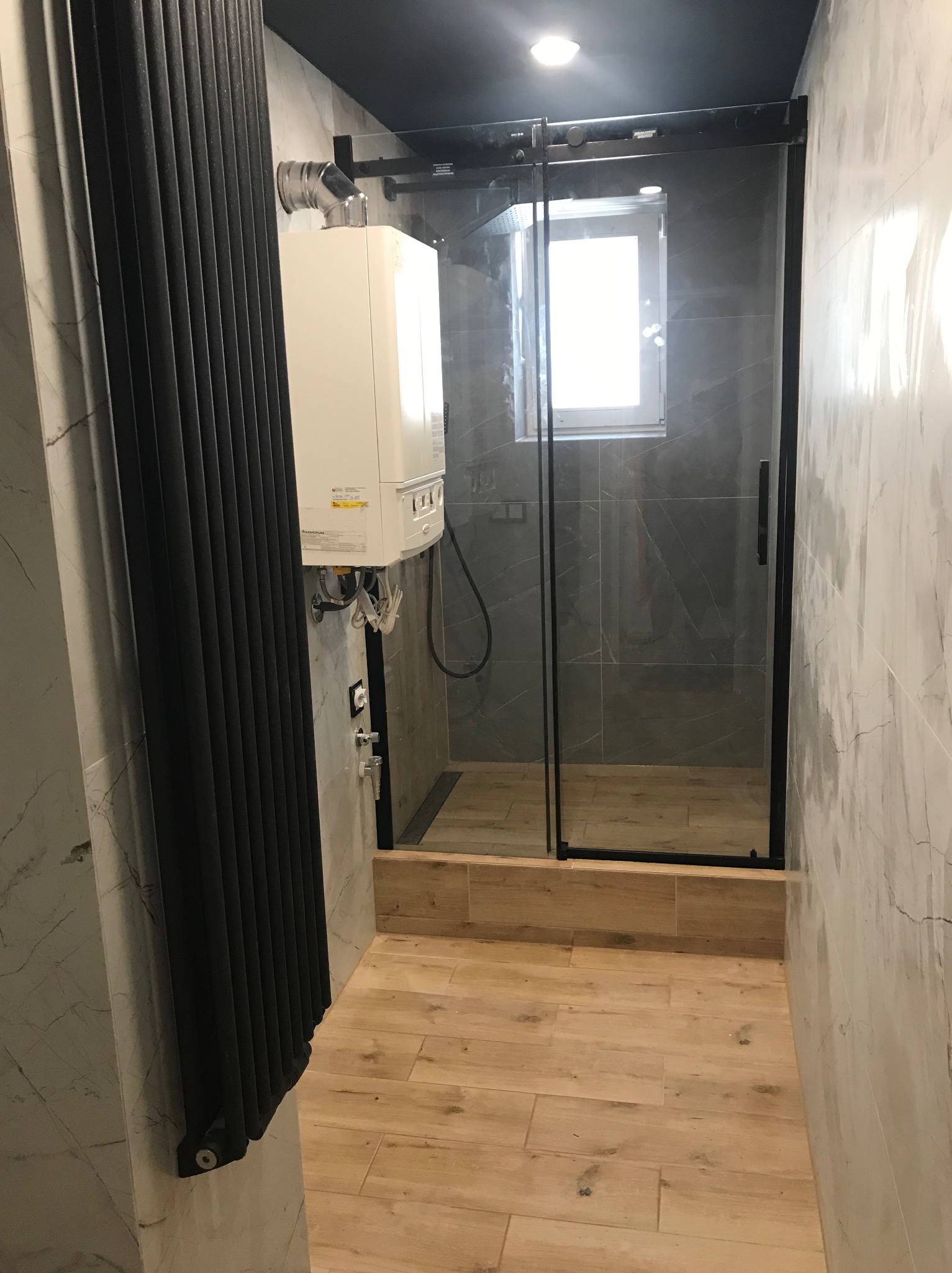 Nowoczesna łazienka z kabiną prysznicową typu walk-in, grzejnikiem żeberkowym i piecem gazowym. Marmurowe płytki ścienne i drewnopodobna podłoga.