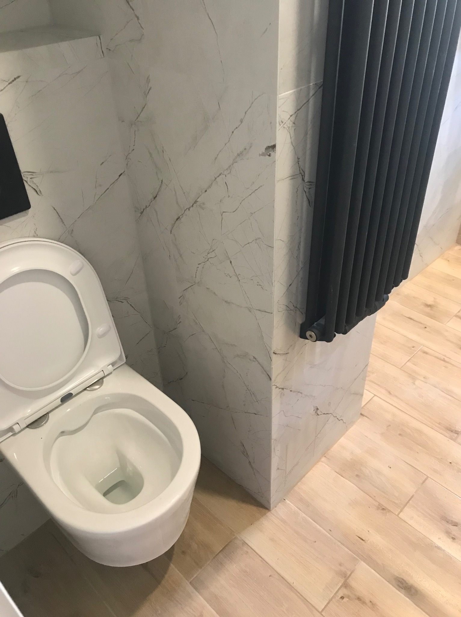 Biała toaleta wisząca z otwartą klapą na tle marmurowych płytek i czarnego grzejnika żeberkowego. Jasne drewnopodobne płytki podłogowe.