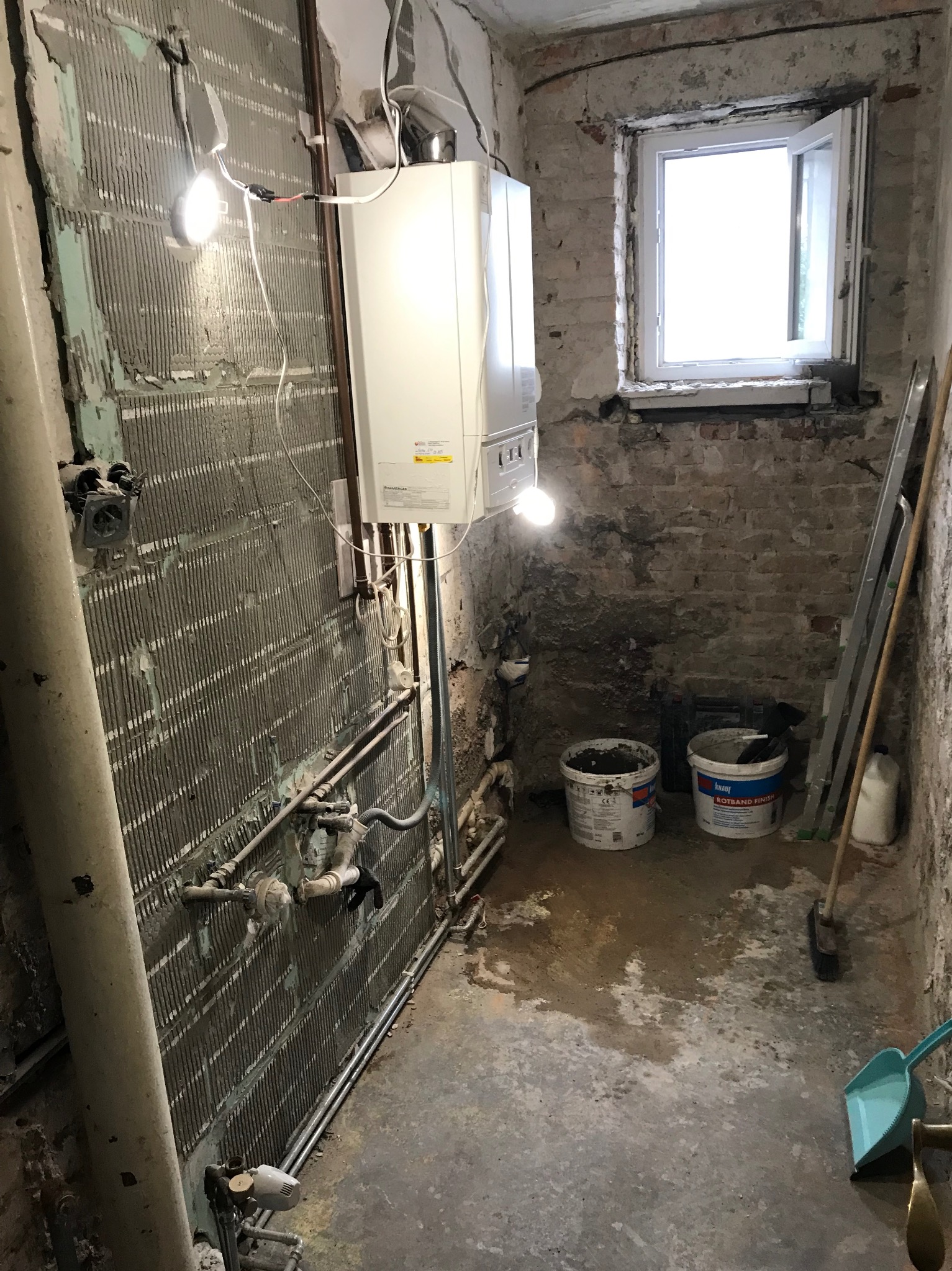 Łazienka w trakcie remontu: ściany z resztkami płytek, odsłonięte rury, piec gazowy, okno, drabina i wiadra z zaprawą. Widoczne ślady wilgoci na podłodze.
