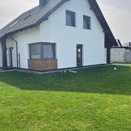 Firma Usługowa Norbert Weber - Biały dom jednorodzinny z ciemną dachówką i drewnianymi akcentami, widoczny z perspektywy ogrodu z zielonym trawnikiem. Świeżo pomalowana elewacja.