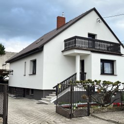 Firma Usługowa Norbert Weber - Biały dom z balkonem i brązowym dachem po renowacji, widoczny z zewnątrz, z otwartą bramą i częściowo widocznym samochodem dostawczym.