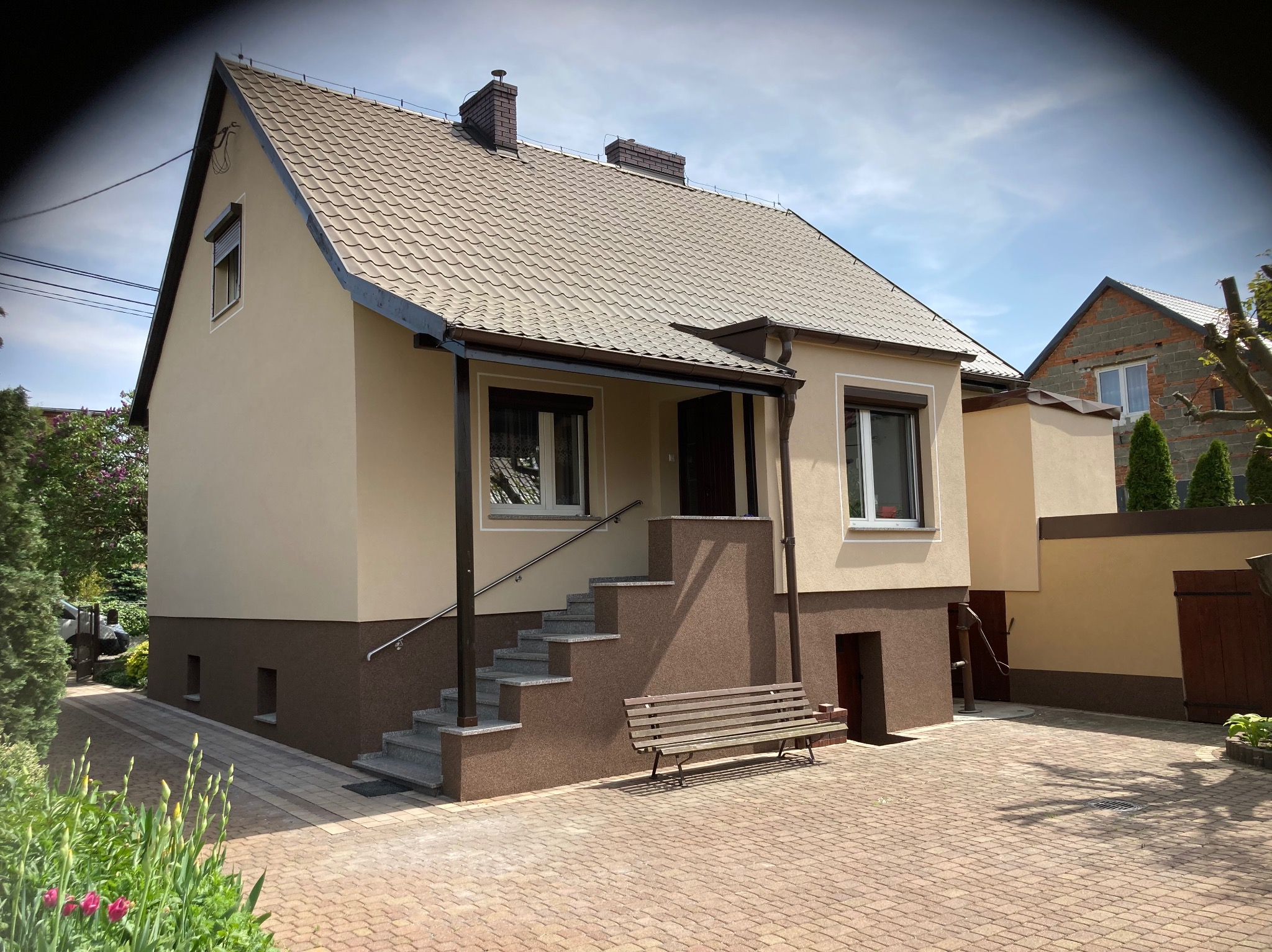 Elewacja domu jednorodzinnego po remoncie, z beżową górą i brązowym dołem, schody wejściowe z ławką, kostka brukowa na podjeździe, w tle budynek w trakcie budowy.