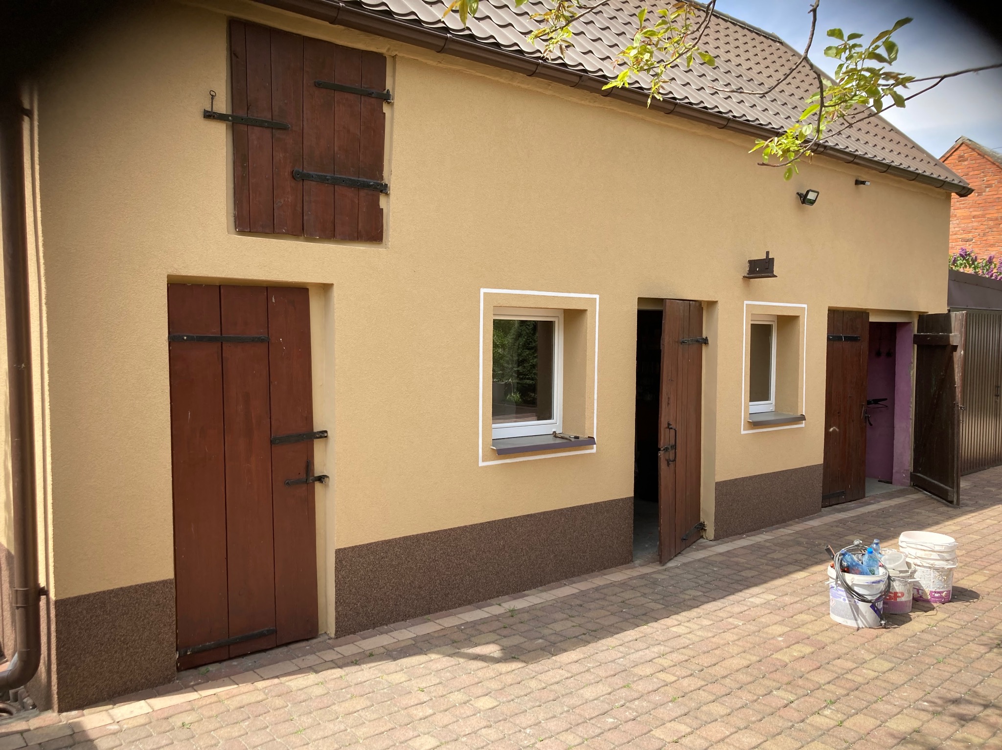 Elewacja budynku po renowacji z drewnianymi okiennicami i drzwiami. Widoczne wiaderka z farbą, sugerujące świeżo zakończone prace malarskie.