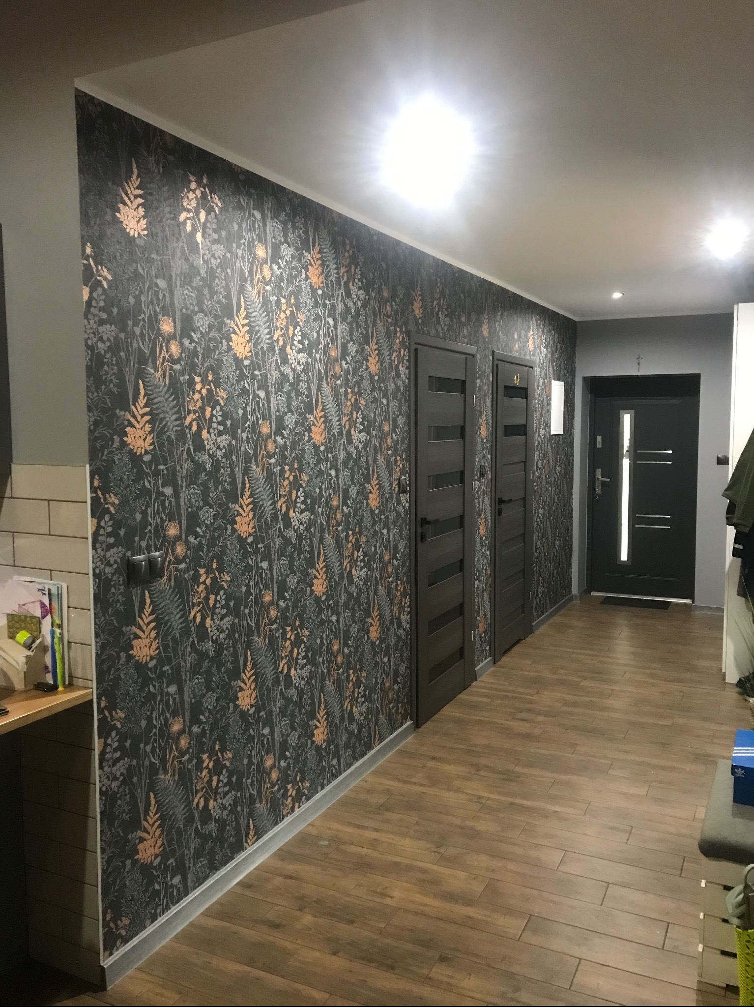 Korytarz po remoncie: wzorzysta tapeta w odcieniach szarości i brązu, drewnopodobne panele podłogowe, dwoje ciemnych drzwi wewnętrznych i drzwi wejściowe.