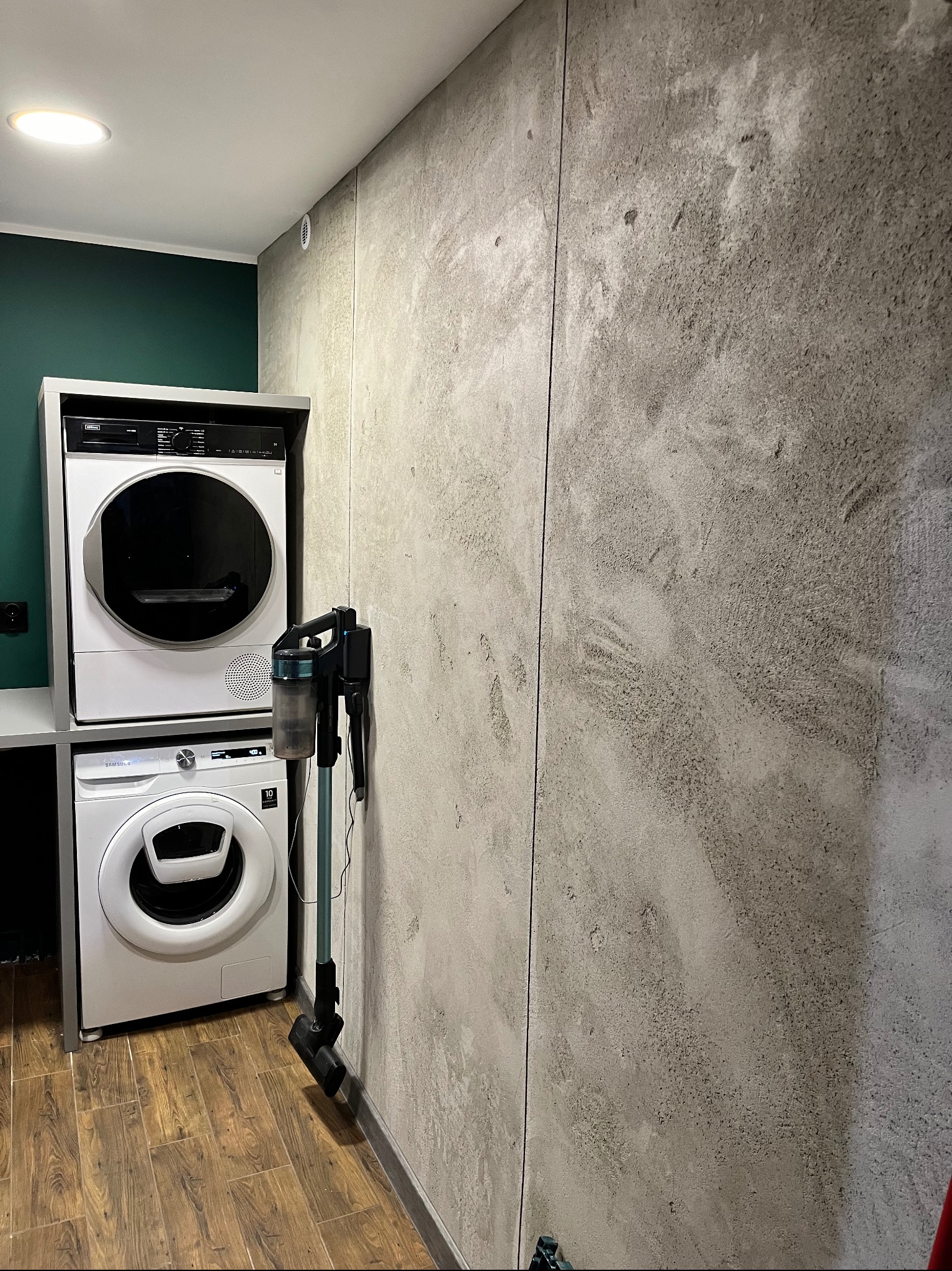 Ściana z efektem betonu architektonicznego w pralni z pralką, suszarką i odkurzaczem pionowym. Wykończenie wnętrza w nowoczesnym stylu.