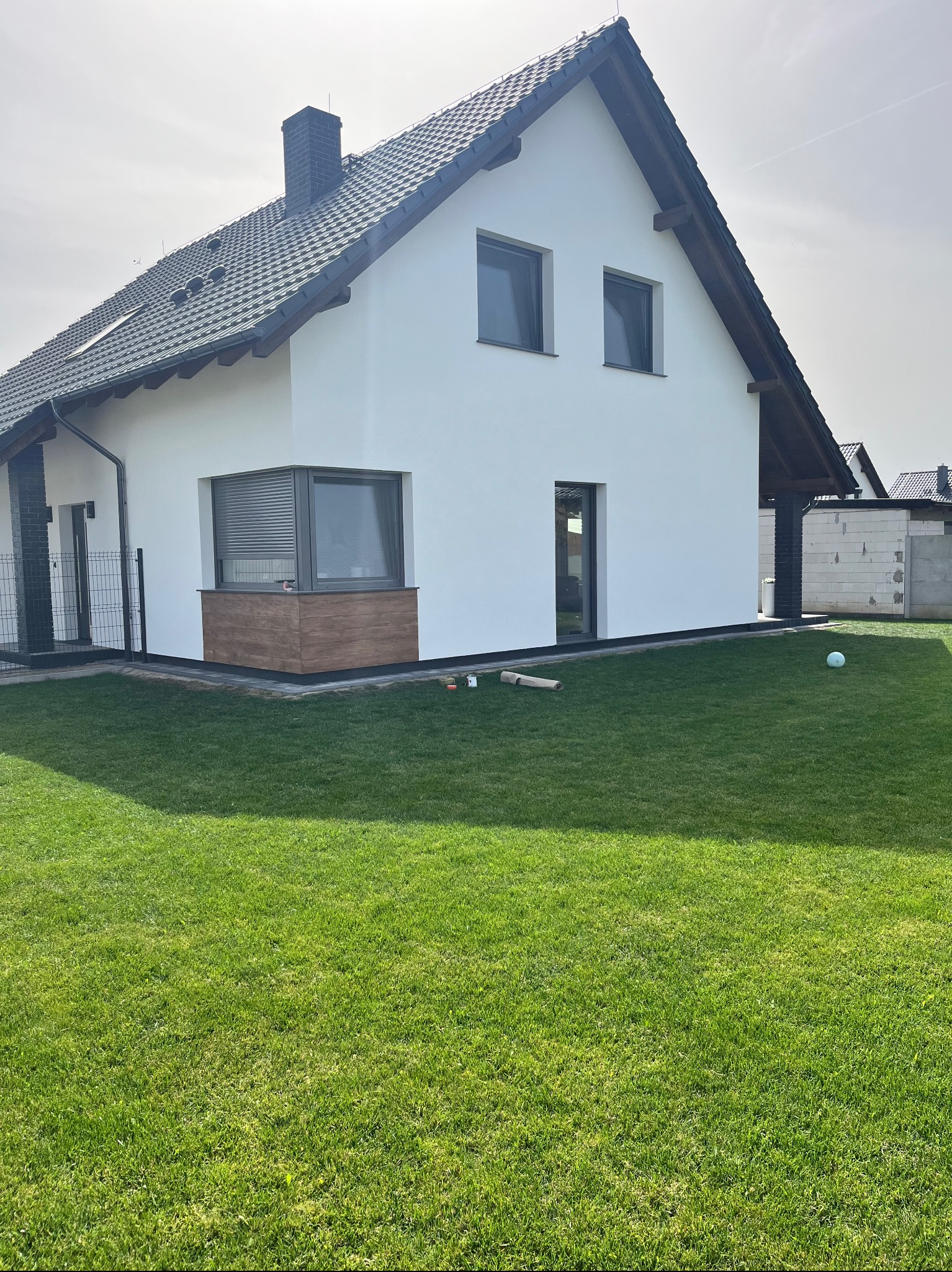 Biały dom jednorodzinny z ciemną dachówką i drewnianymi akcentami, widoczny z perspektywy ogrodu z zielonym trawnikiem. Świeżo pomalowana elewacja.