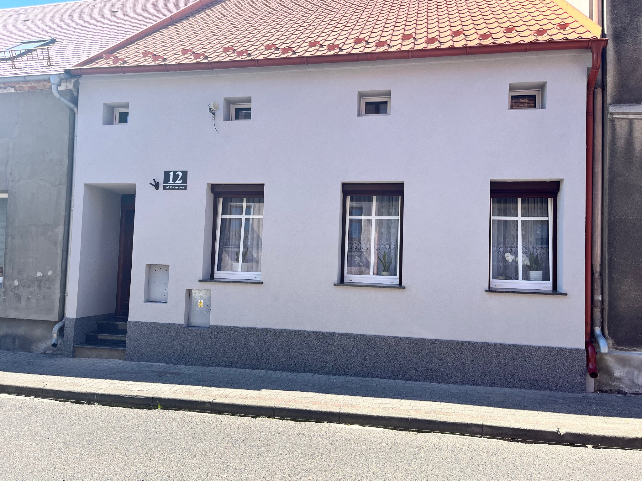Odnowiona fasada domu z numerem 12, z nową elewacją w odcieniach szarości i białym tynkiem. Widoczne okna z roletami i czerwony dach.