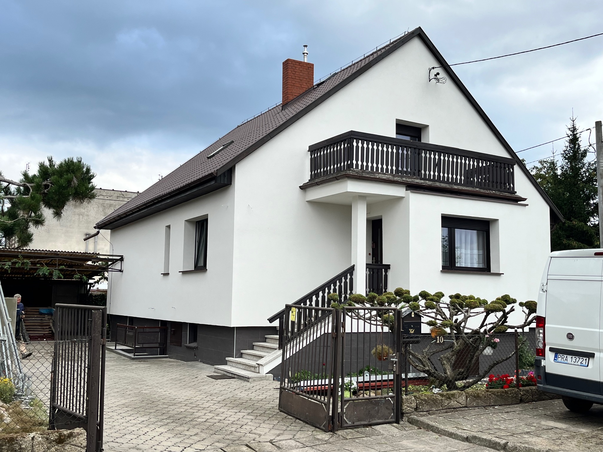 Biały dom z balkonem i brązowym dachem po renowacji, widoczny z zewnątrz, z otwartą bramą i częściowo widocznym samochodem dostawczym.